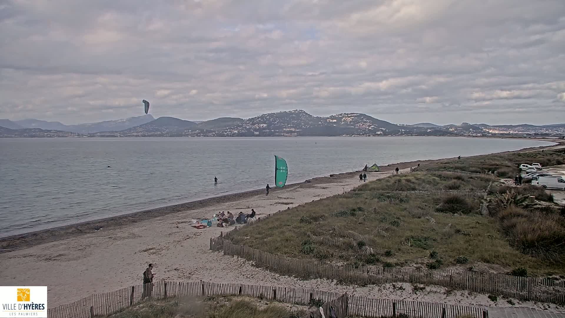 A sandy beach with people relaxing and kitesurfing is seen under a cloudy sky, with a calm body of water and distant mountains in the background.