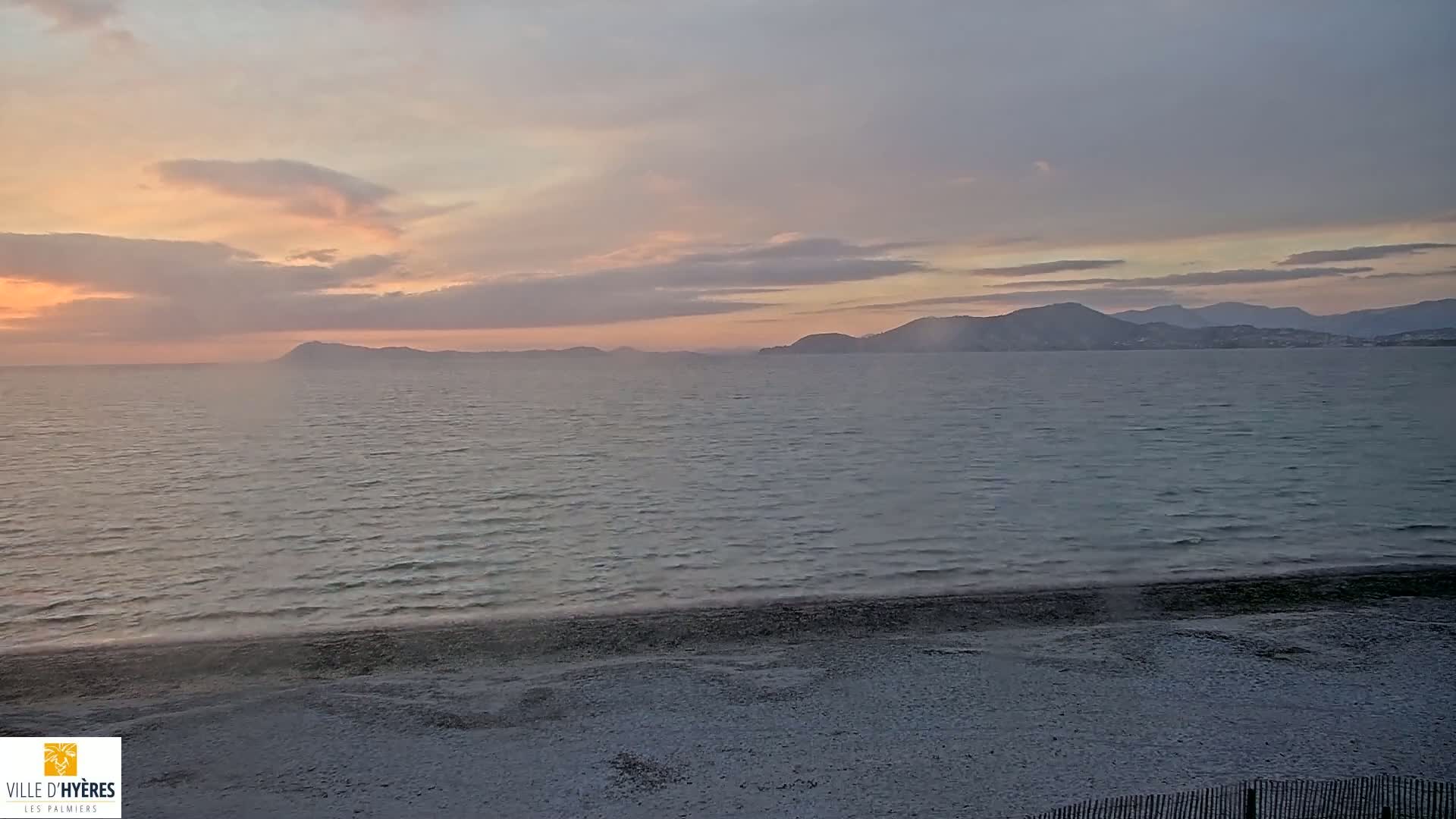 A calm sea with small waves laps a sandy beach under a pale sunset sky with distant mountains.