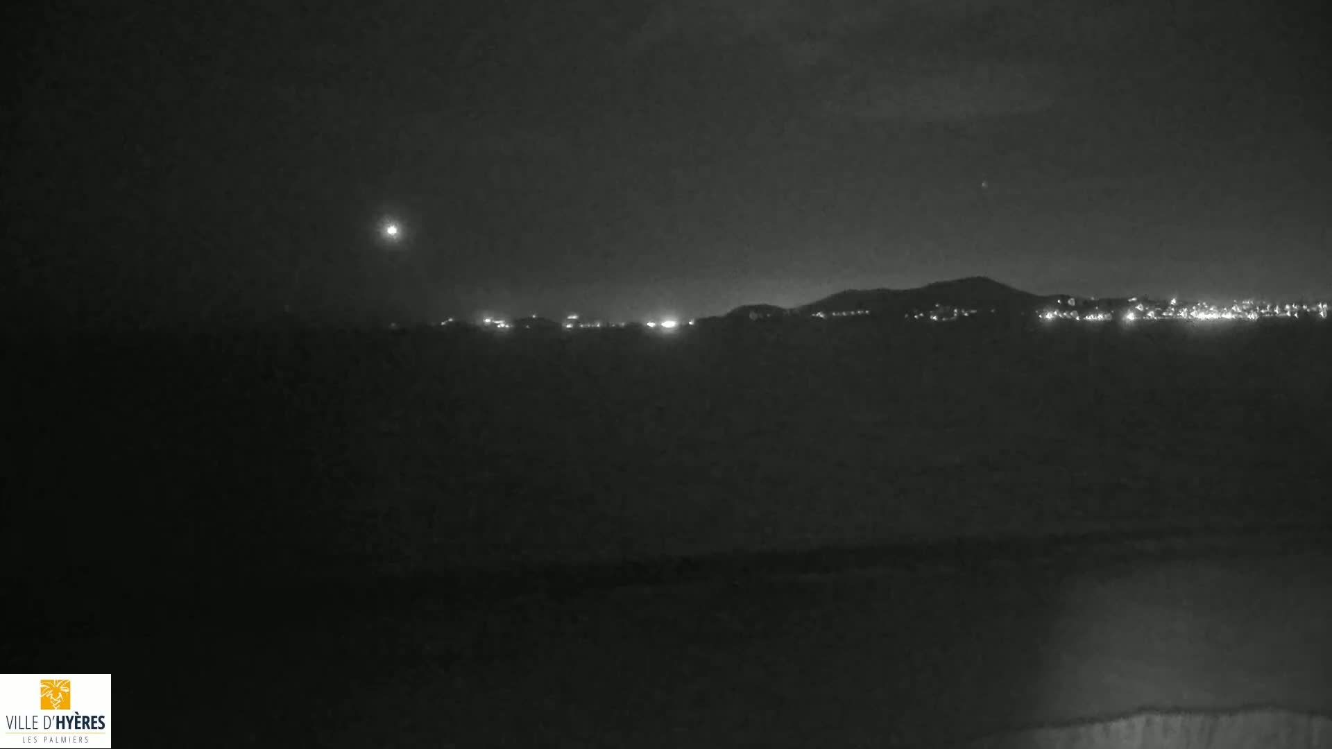 A nighttime, low-light image shows a dark, hilly landscape with a line of lights on the horizon under a mostly clear sky.