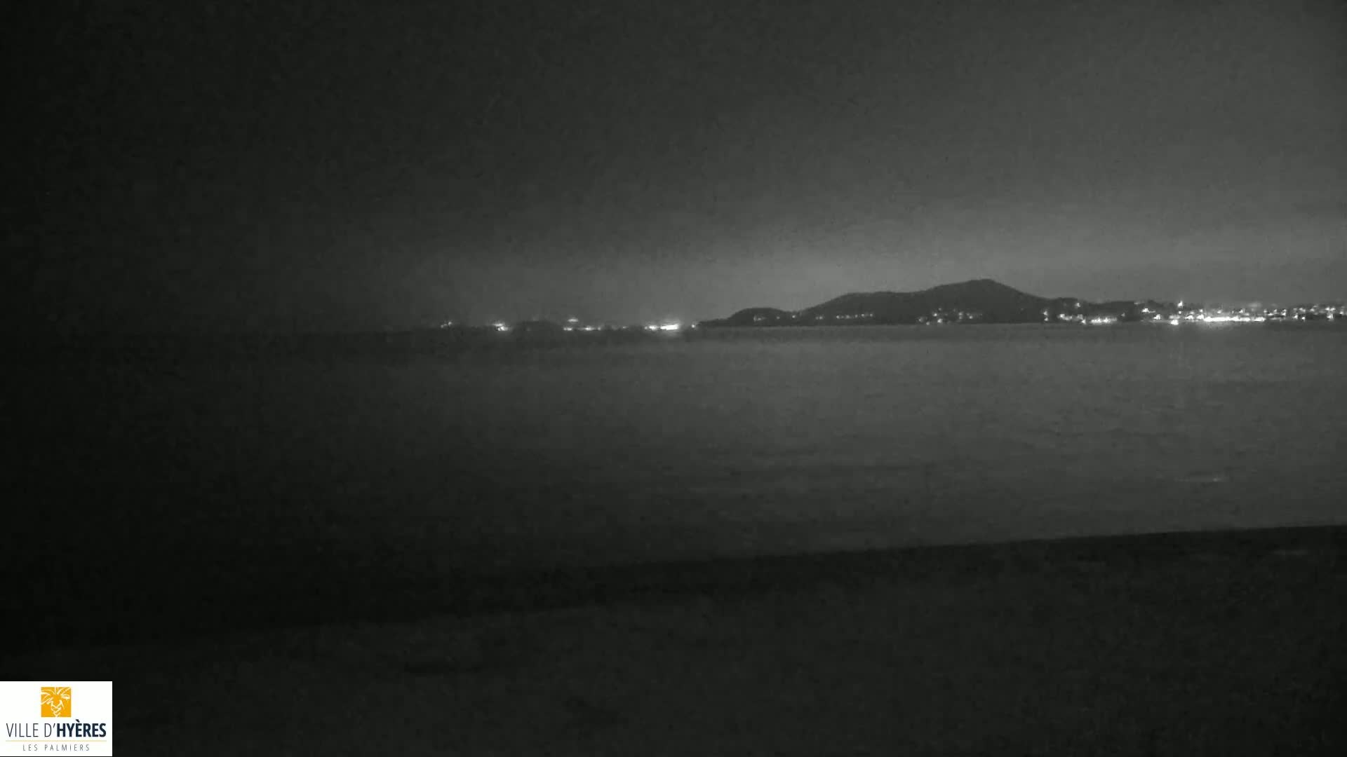 A nighttime, dark grayscale image shows a body of water with a distant shoreline featuring city lights and hills under a cloudy sky.