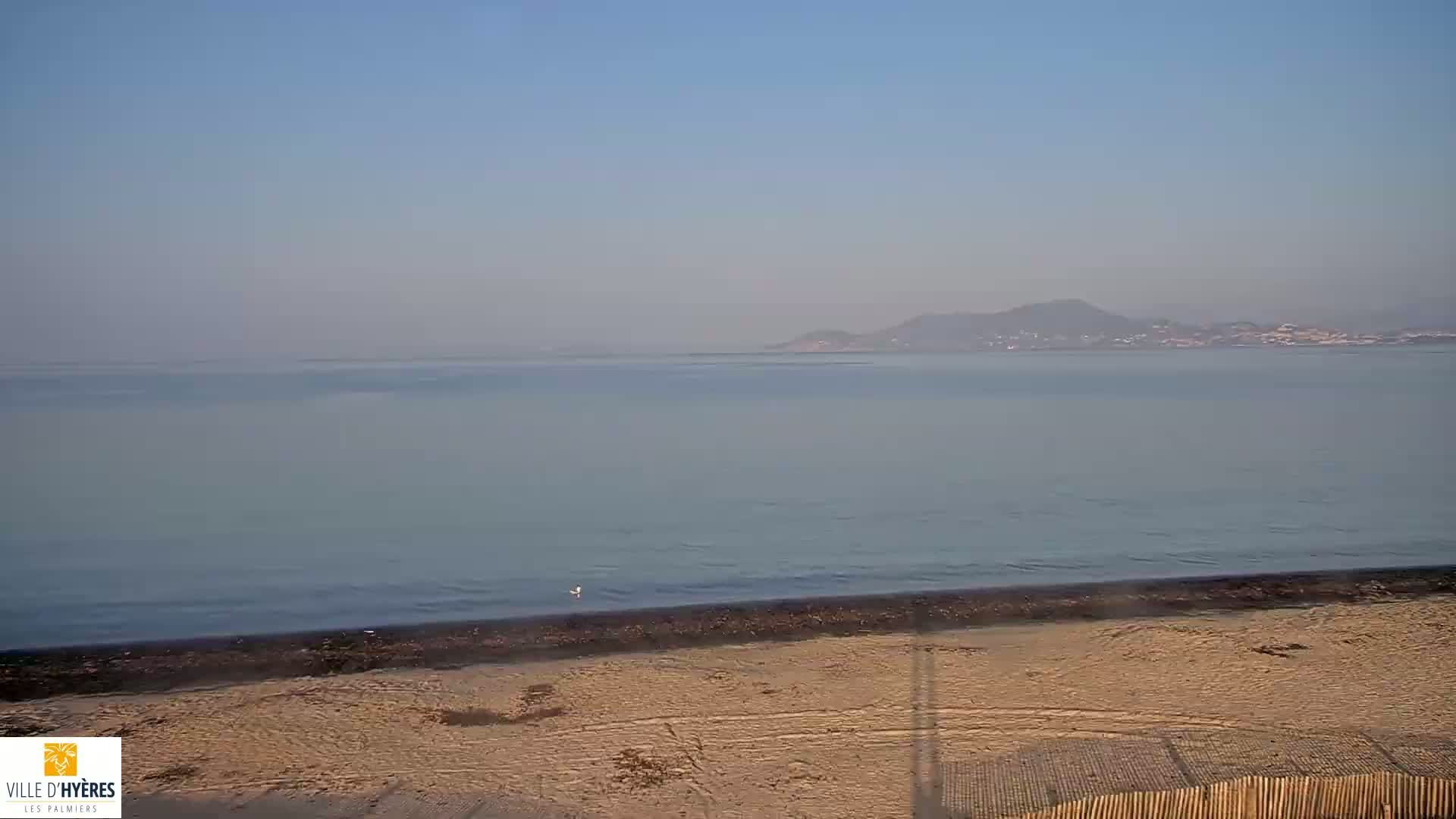 A hazy, calm ocean meets a sandy beach backed by a low, distant mountain range.