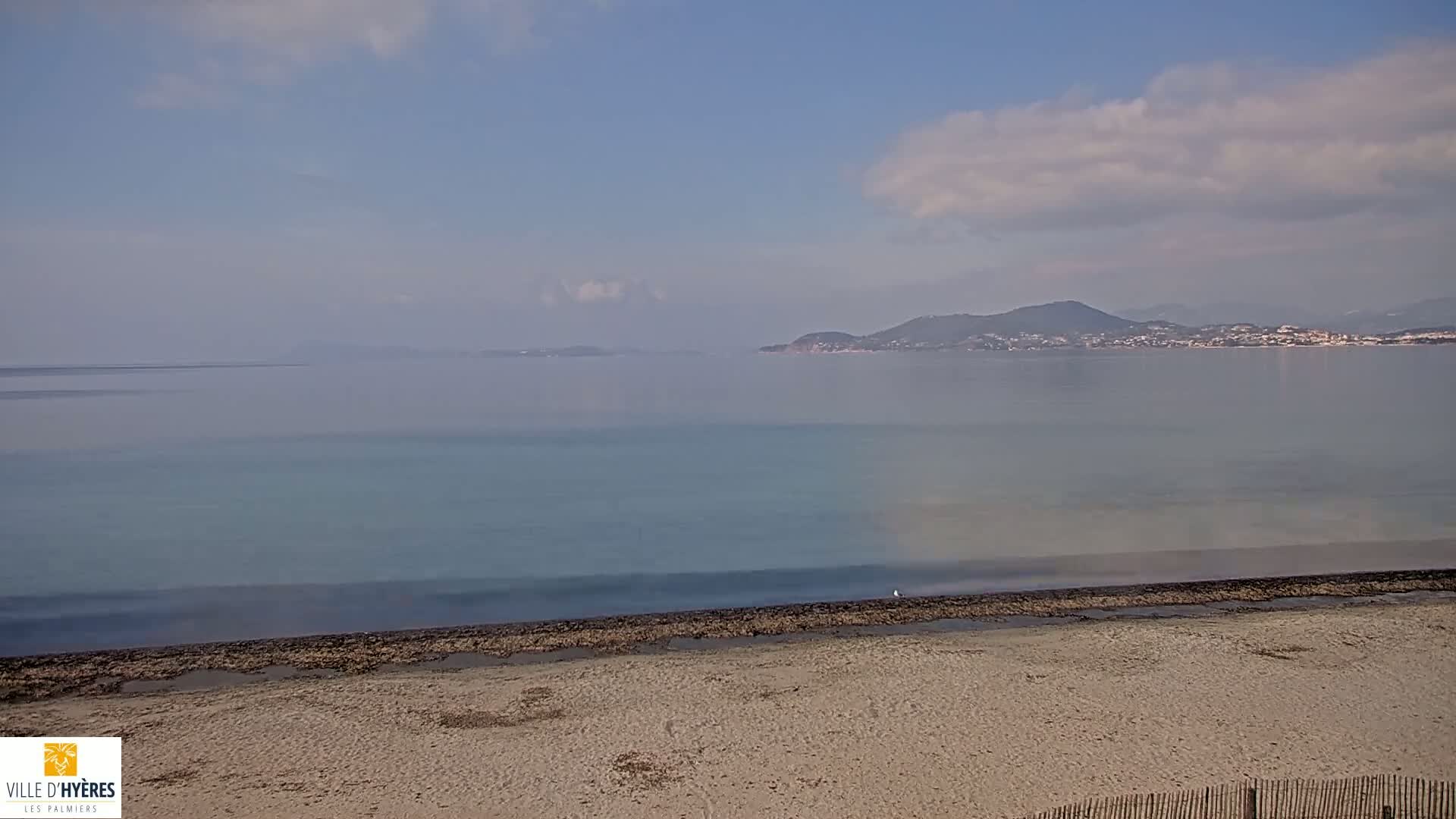 A hazy, partly cloudy day reveals a sandy beach leading to a calm, pale-blue sea, with distant landmasses visible on the horizon.