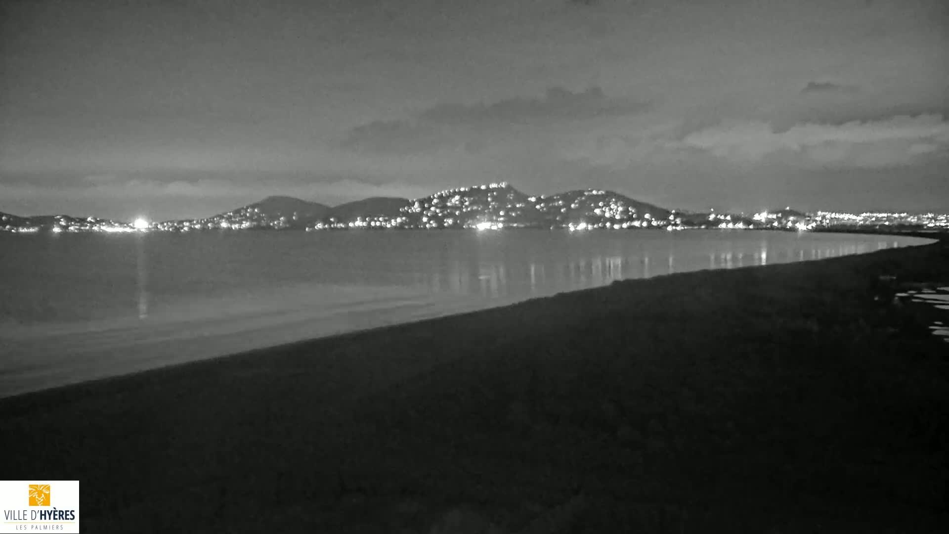 A dark, nighttime beach scene shows a calm body of water reflecting the lights of a distant city nestled in hills under a cloudy sky.