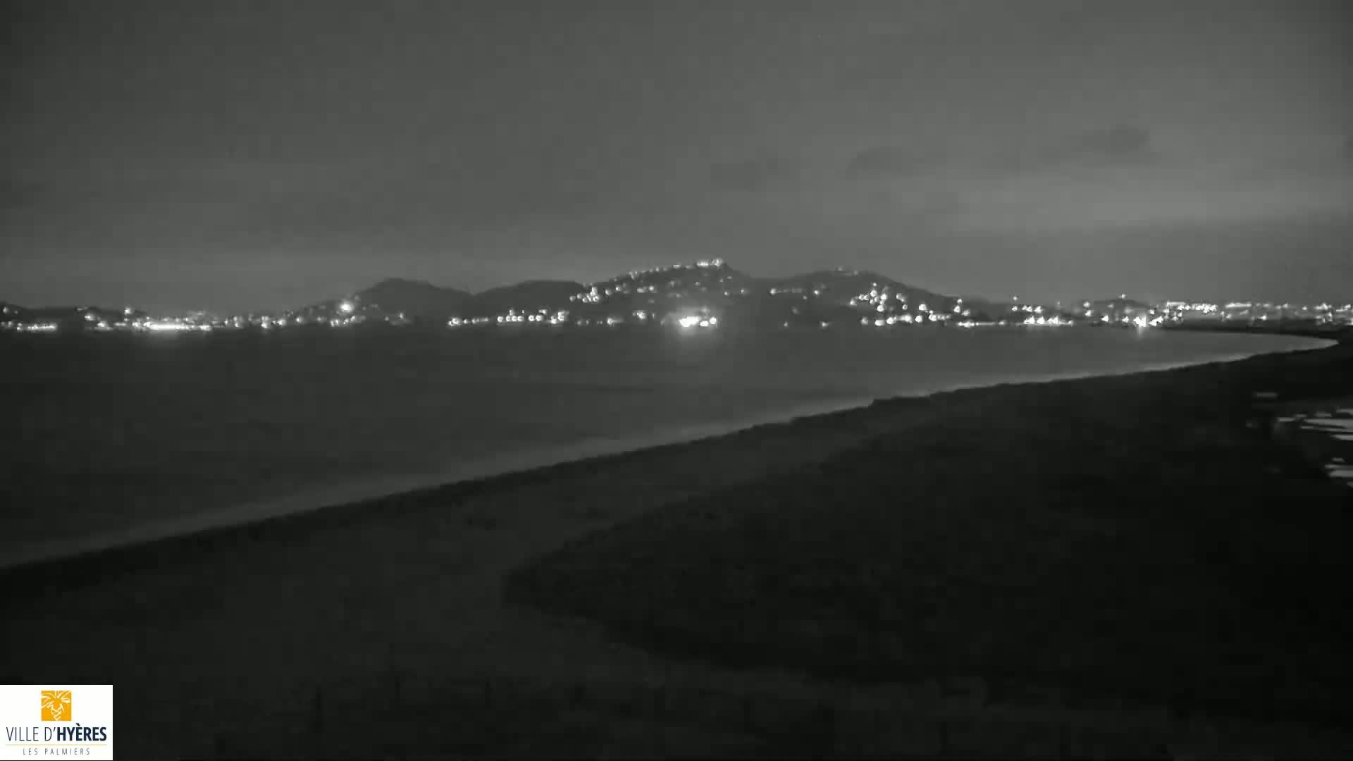 A nighttime, grayscale view shows a dark coastline with a distant, hilly town illuminated by lights under a cloudy sky.