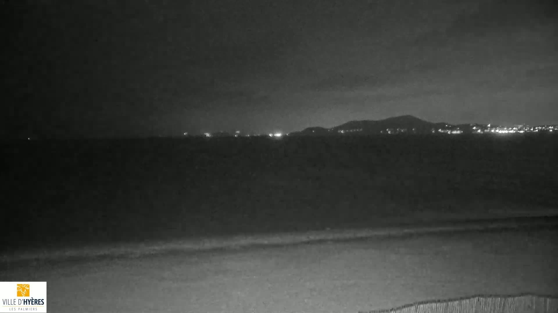 A dark, nighttime view of a calm body of water with a distant, dimly lit shoreline and hills under a cloudy sky.