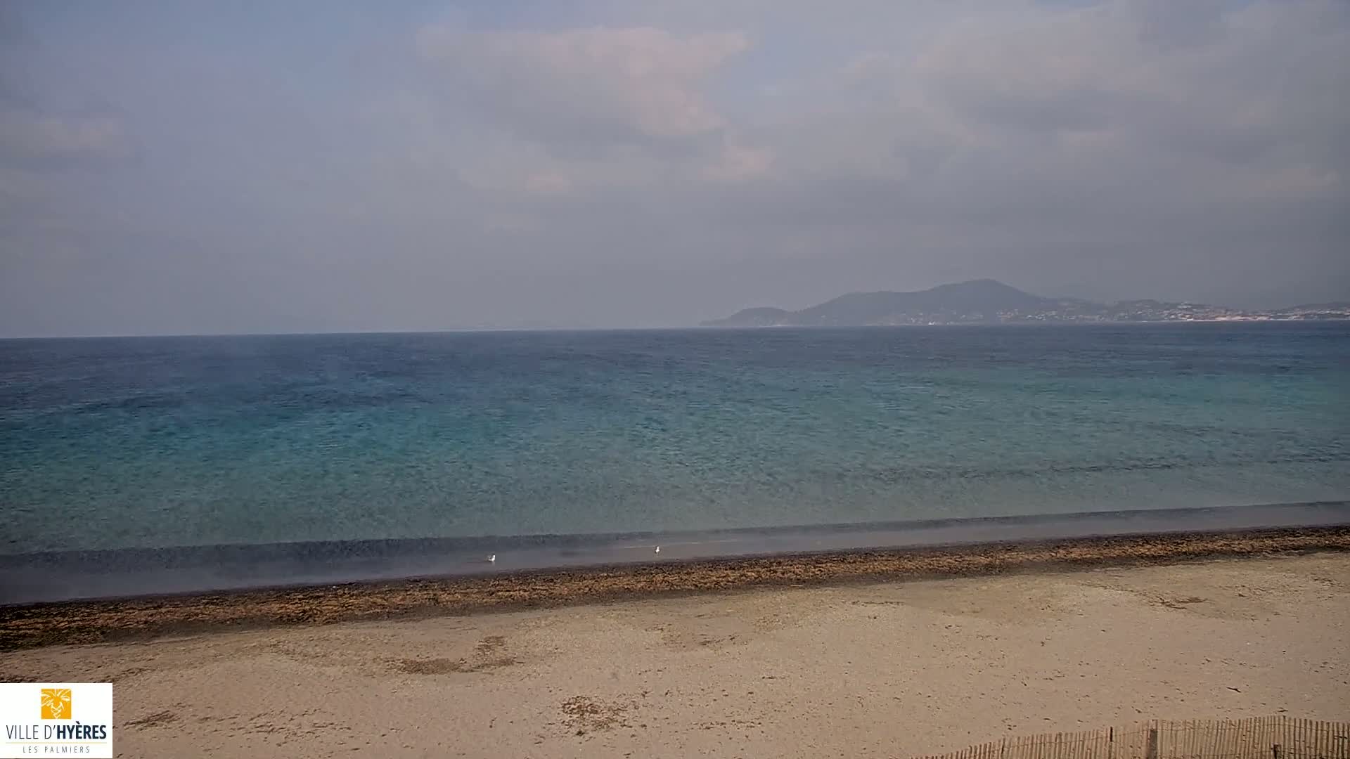 A sandy beach stretches to a calm, teal ocean under a hazy sky, with a distant mountain visible on the horizon.