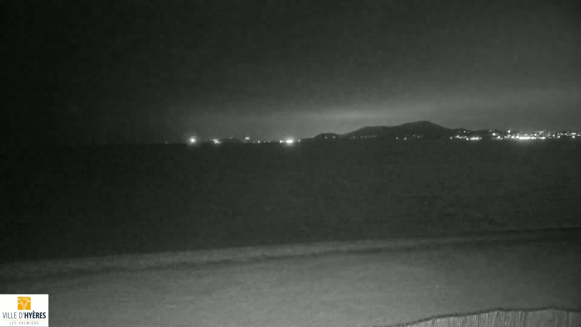A nighttime view of a dark body of water with dimly lit buildings and hills in the distance under a cloudy sky.