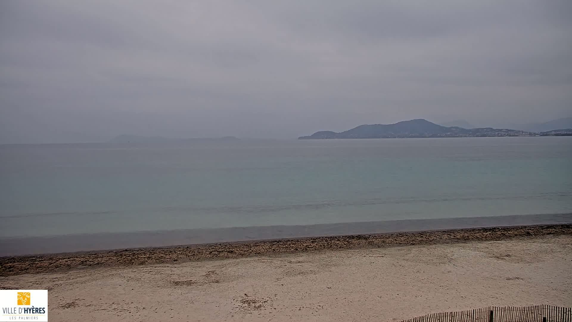 A sandy beach stretches to a calm, light-teal sea under an overcast gray sky, with distant hills visible on the horizon.