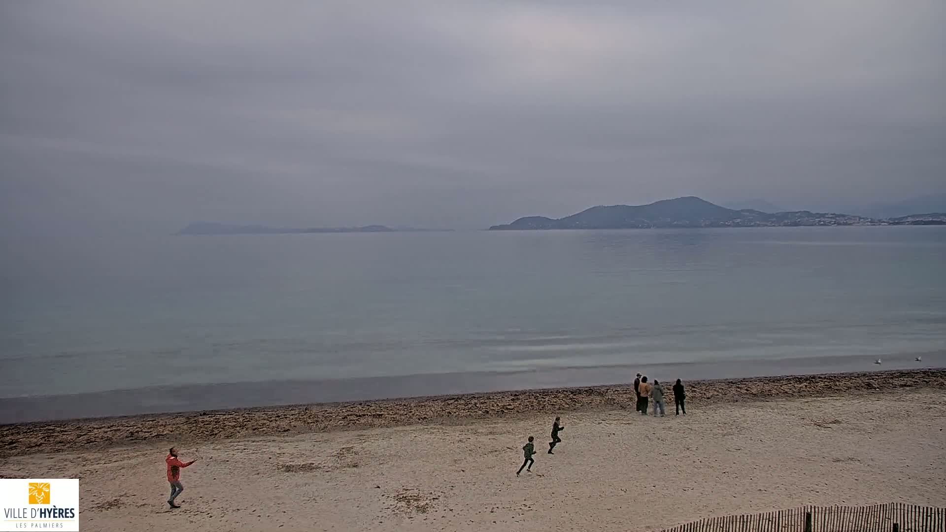 A sandy beach with several people, some running and others standing, overlooks a calm, gray sea under an overcast sky with distant hills visible on the horizon.