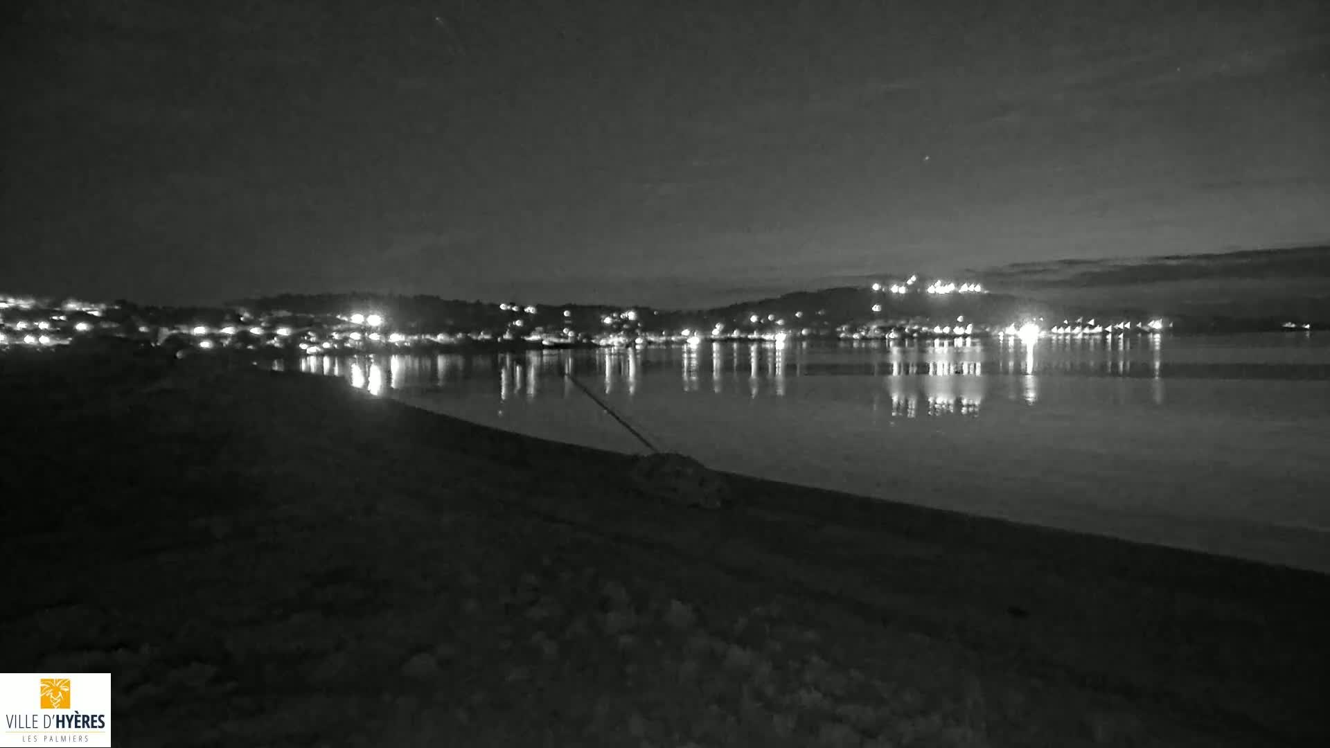 A dark beach at night shows the lights of a town across a calm body of water under a mostly clear sky.