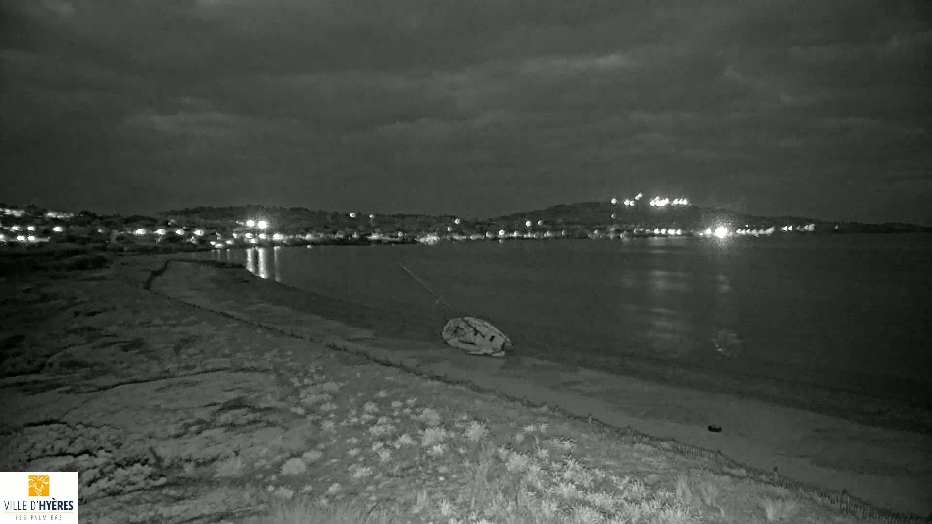 A nighttime, grayscale image shows a dark, calm body of water reflecting the lights of a distant town under a cloudy sky, with a small object lying on the beach in the foreground.