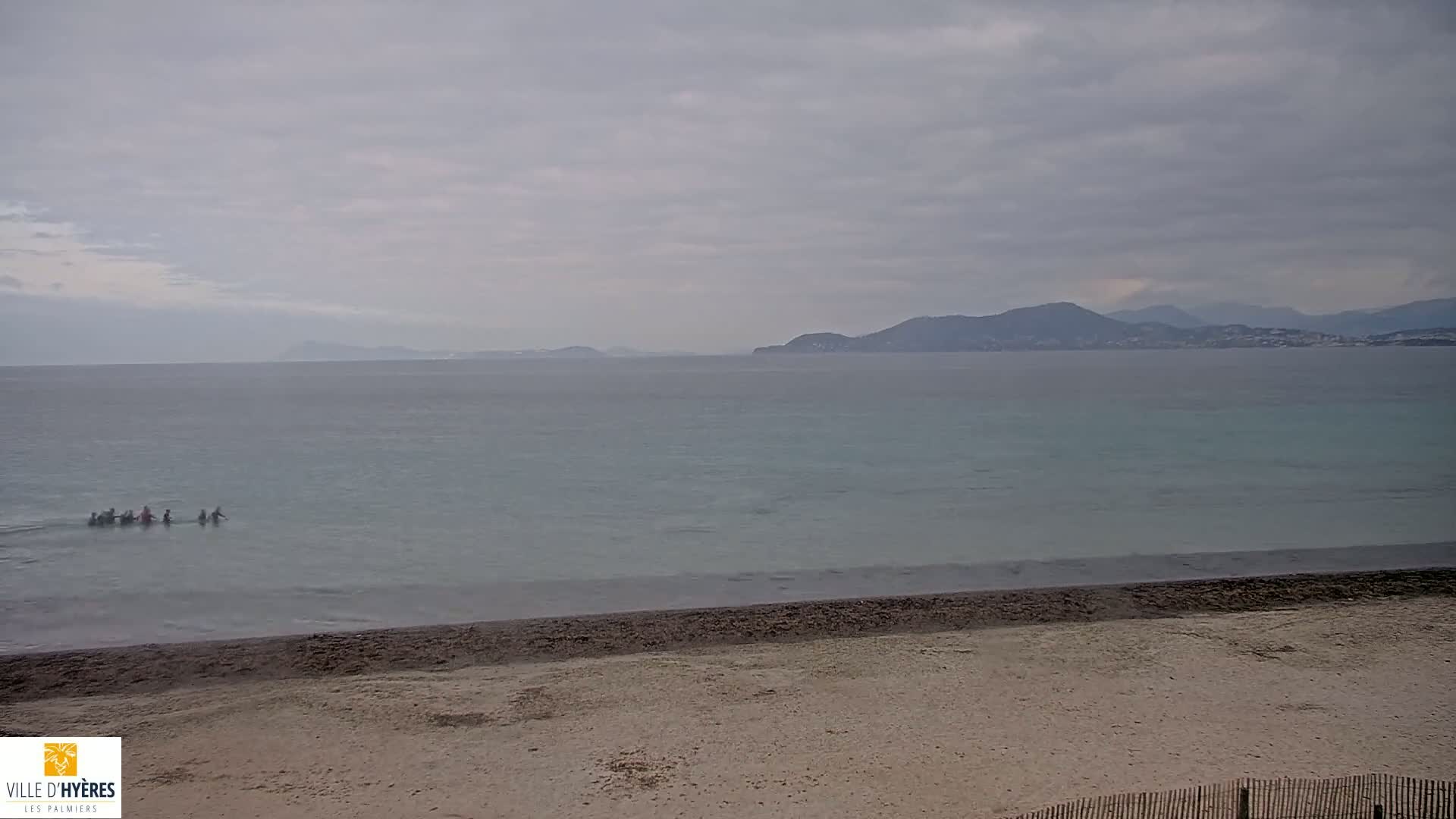 A group of people are wading in calm, pale-blue water near a sandy beach, with distant mountains visible under an overcast sky.