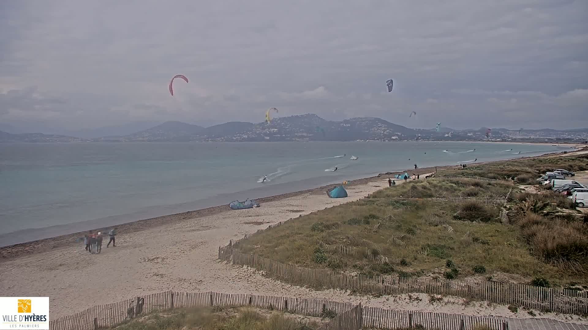 Several kiteboarders are visible on a beach with calm, light-blue water under an overcast sky, with distant hills in the background.