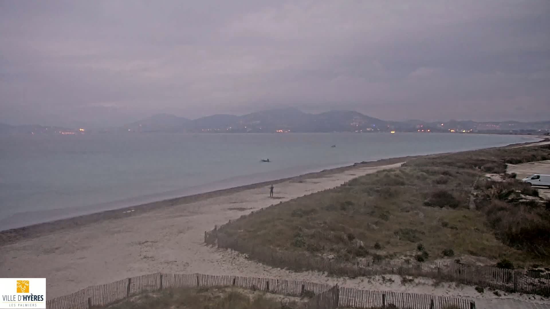 A sandy beach stretches along a calm, gray sea under an overcast sky, backed by low-lying hills with a few distant lights.