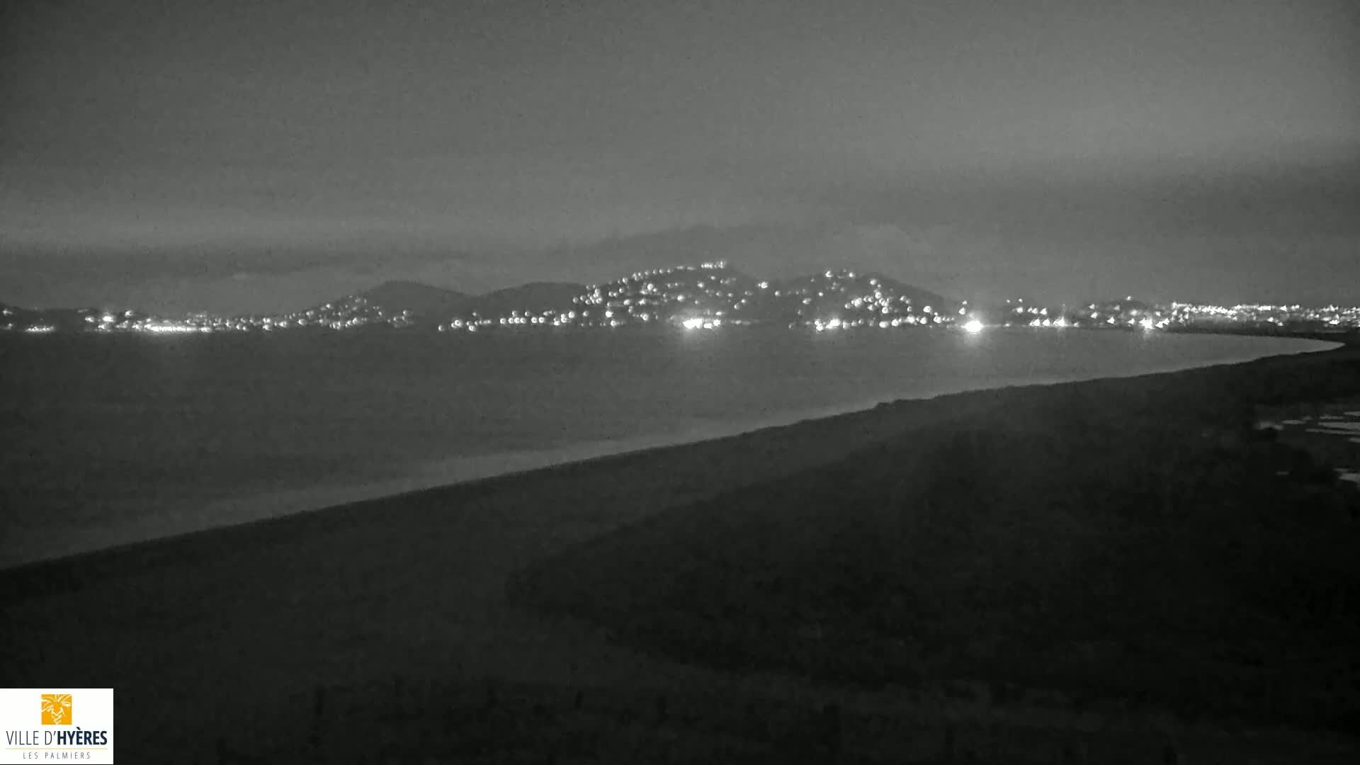 A nighttime, grayscale view shows a dark, calm body of water separating a dark shoreline from a hill dotted with lights in the distance.