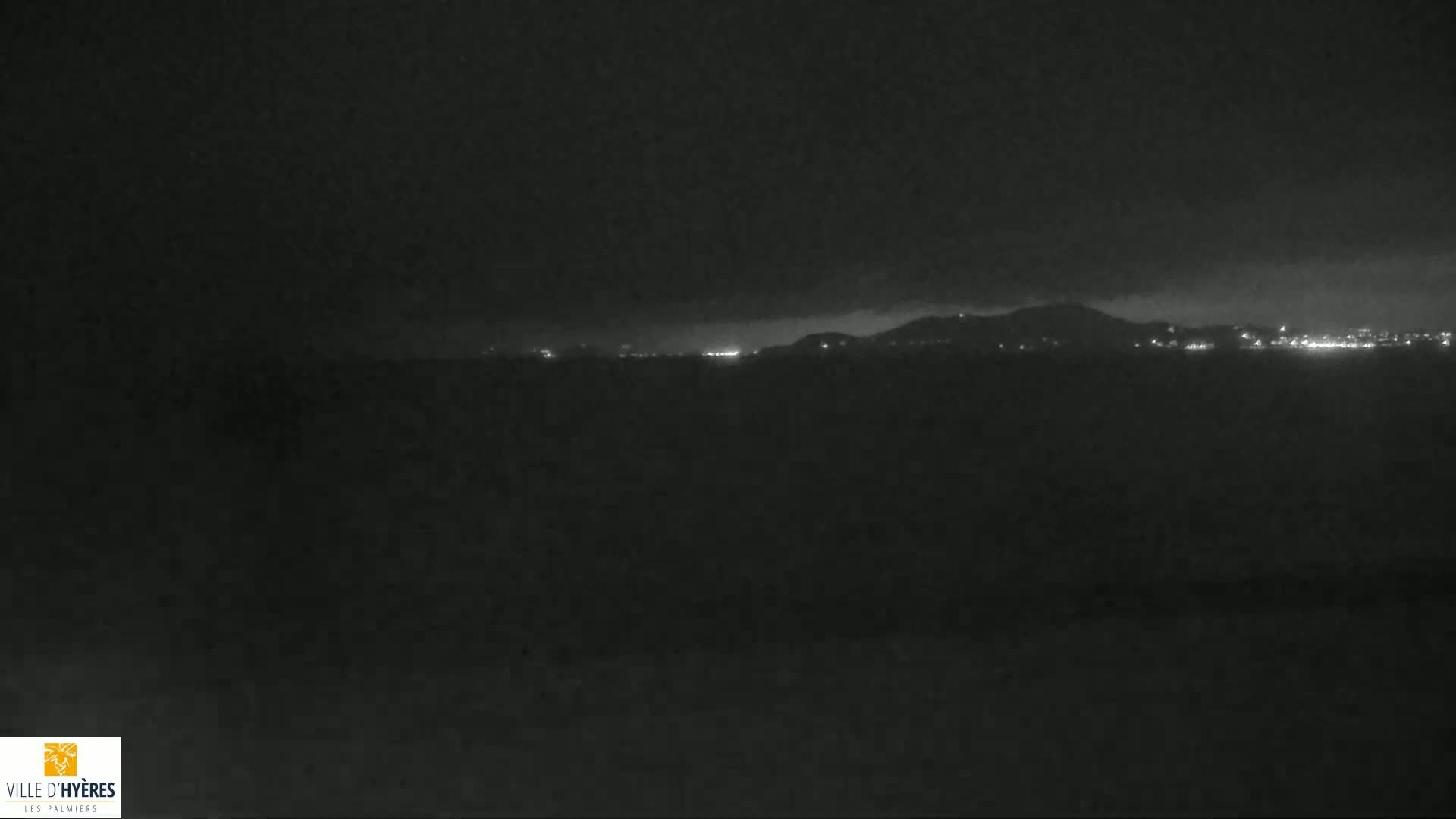 A dark, nighttime scene shows a dimly lit cityscape and hills in the distance under a cloudy sky.