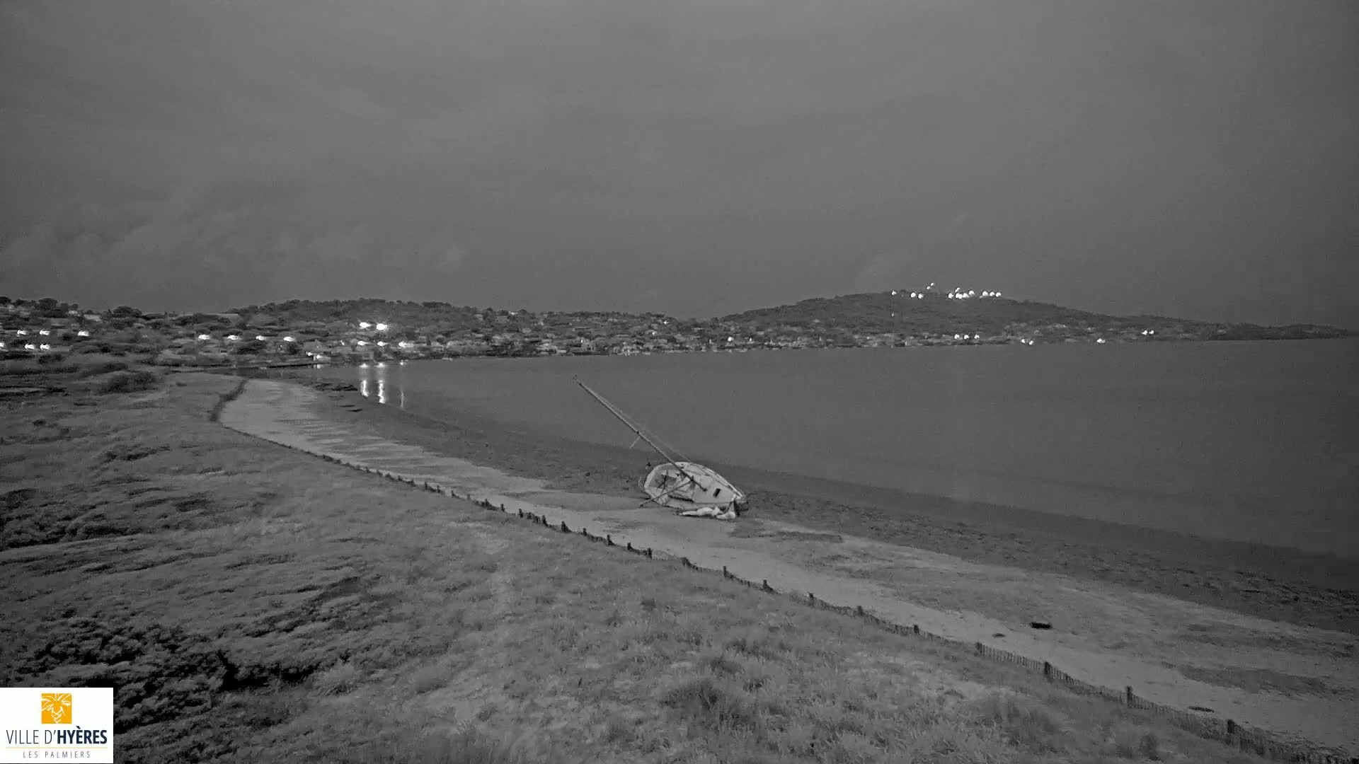A capsized sailboat lies on a beach at dusk, with a town and hill visible across a calm body of water.