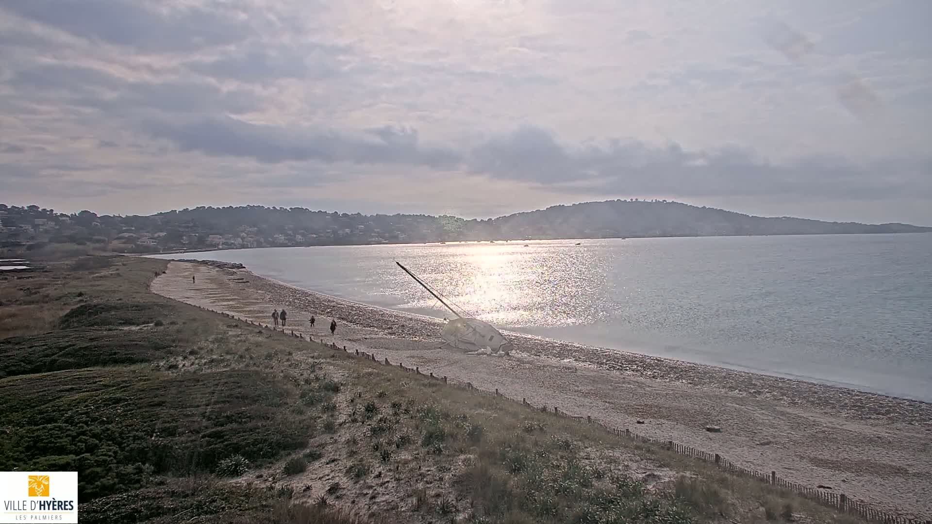 A partly cloudy day shows a sandy beach with a capsized sailboat near a shoreline with a few people walking, and calm water reflecting sunlight in the background.