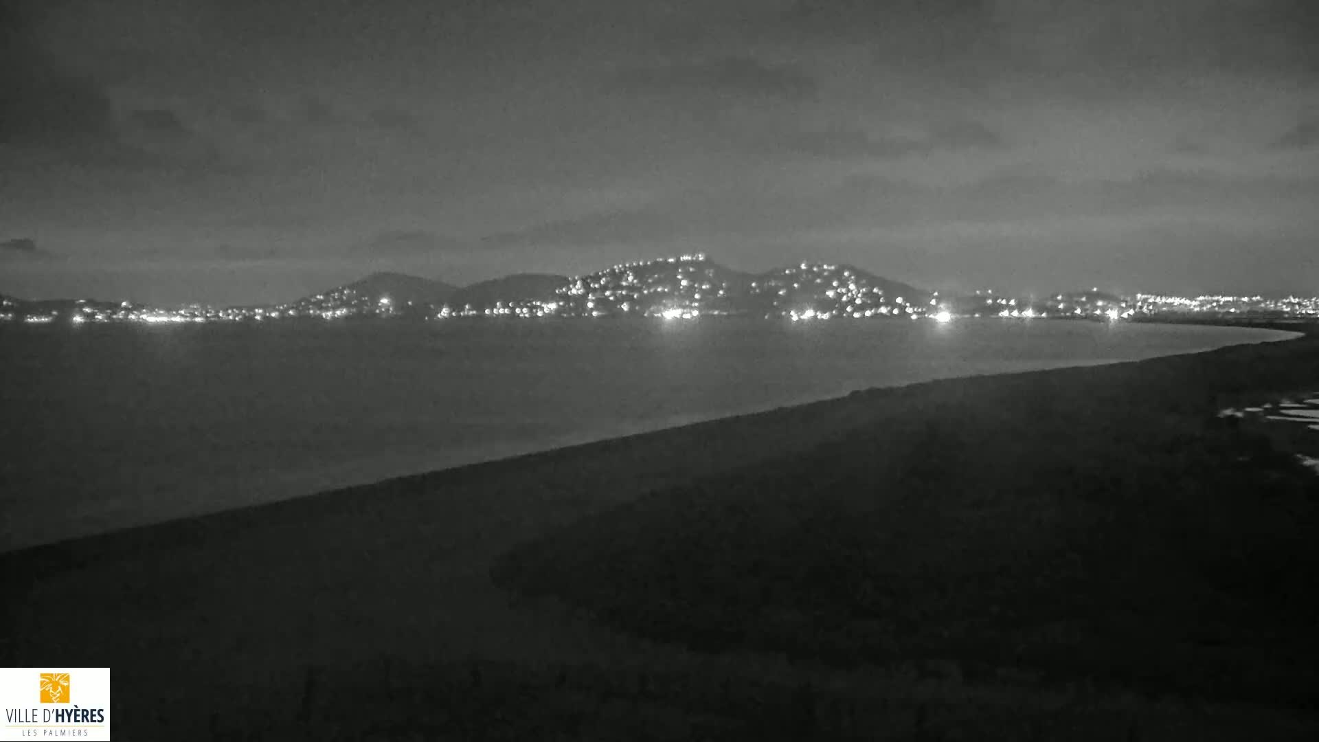 A nighttime, grayscale view shows a city's lights illuminating hills across a dark body of water from a dark, land-based vantage point.