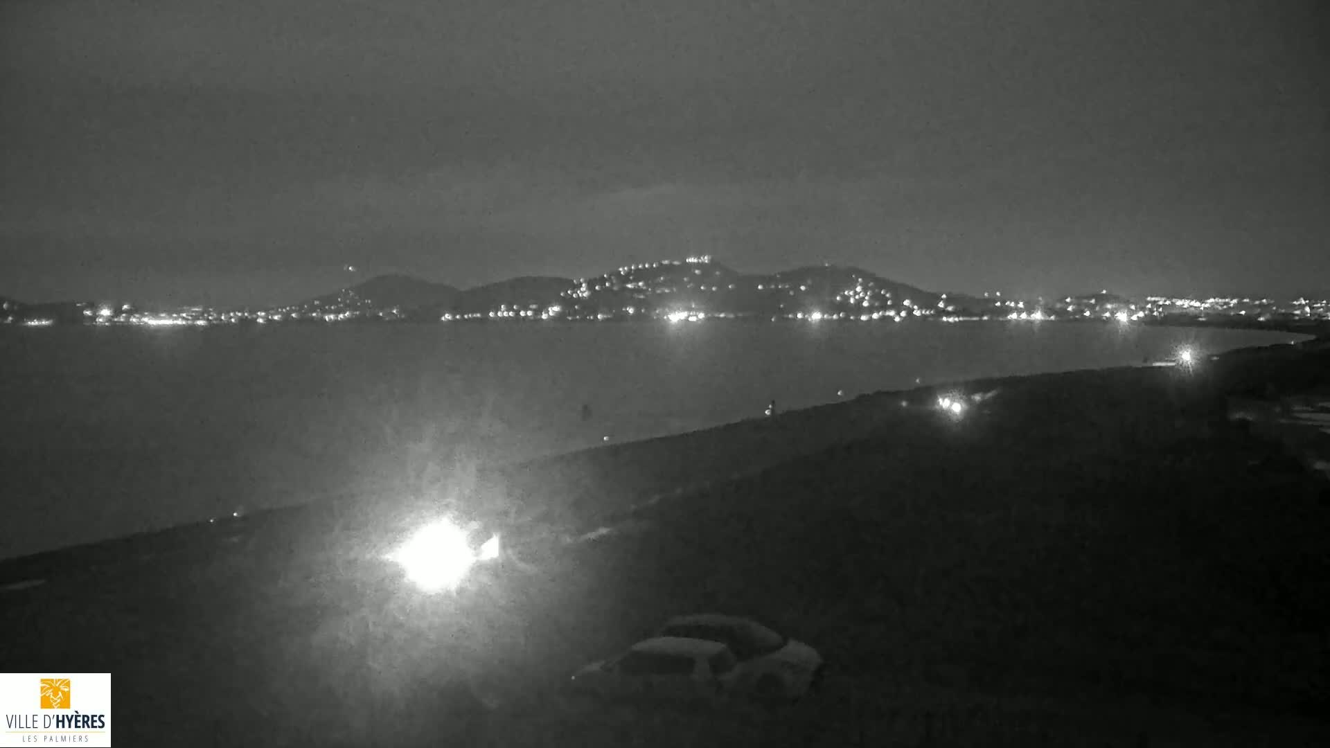 A nighttime view of a city's lights across a body of water, with a few vehicles visible in the foreground under a dark sky.