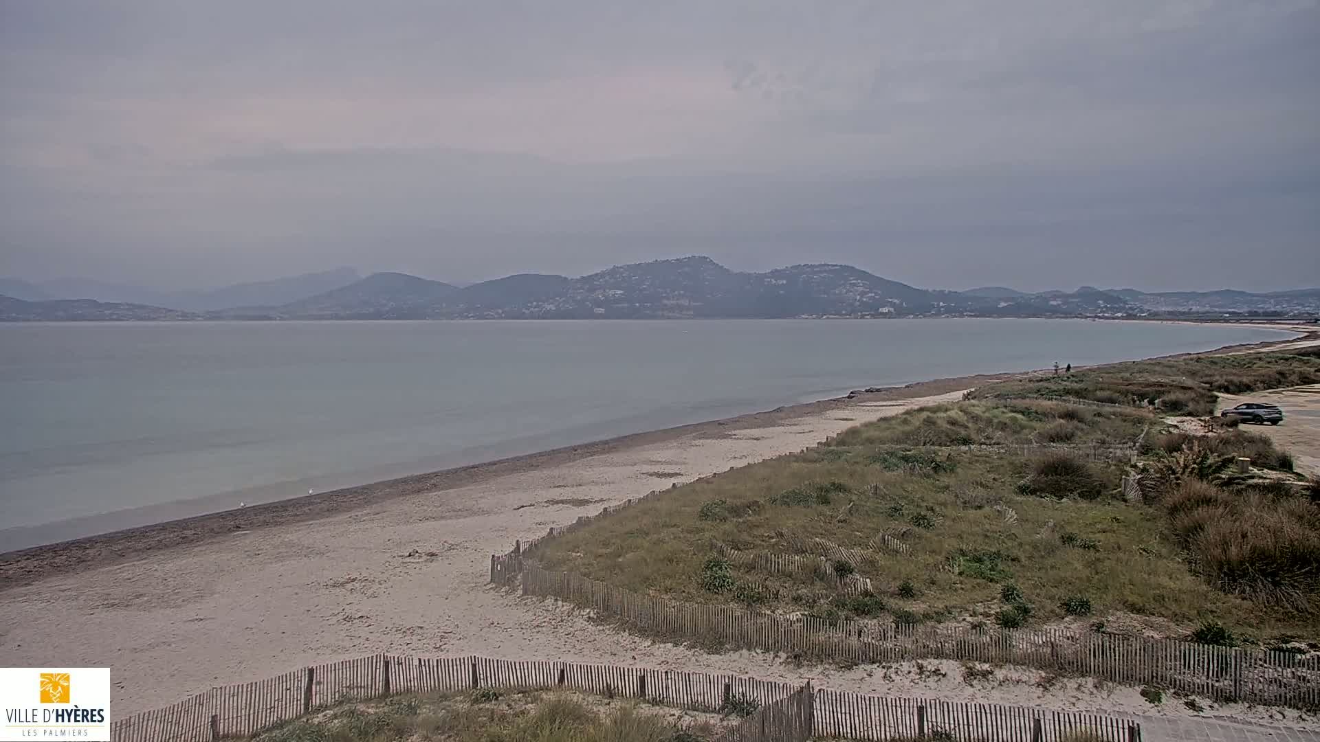 A sandy beach stretches along a calm, light-grey body of water under an overcast sky, with low-lying hills visible in the distance.