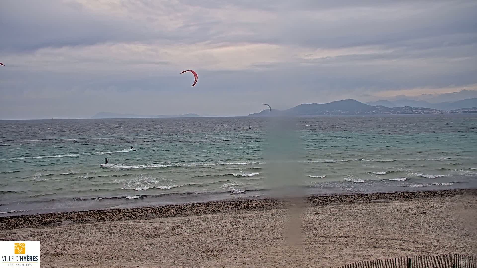 Several kitesurfers are enjoying a windy day on a sandy beach with light waves and a cloudy sky, mountains visible in the distance.