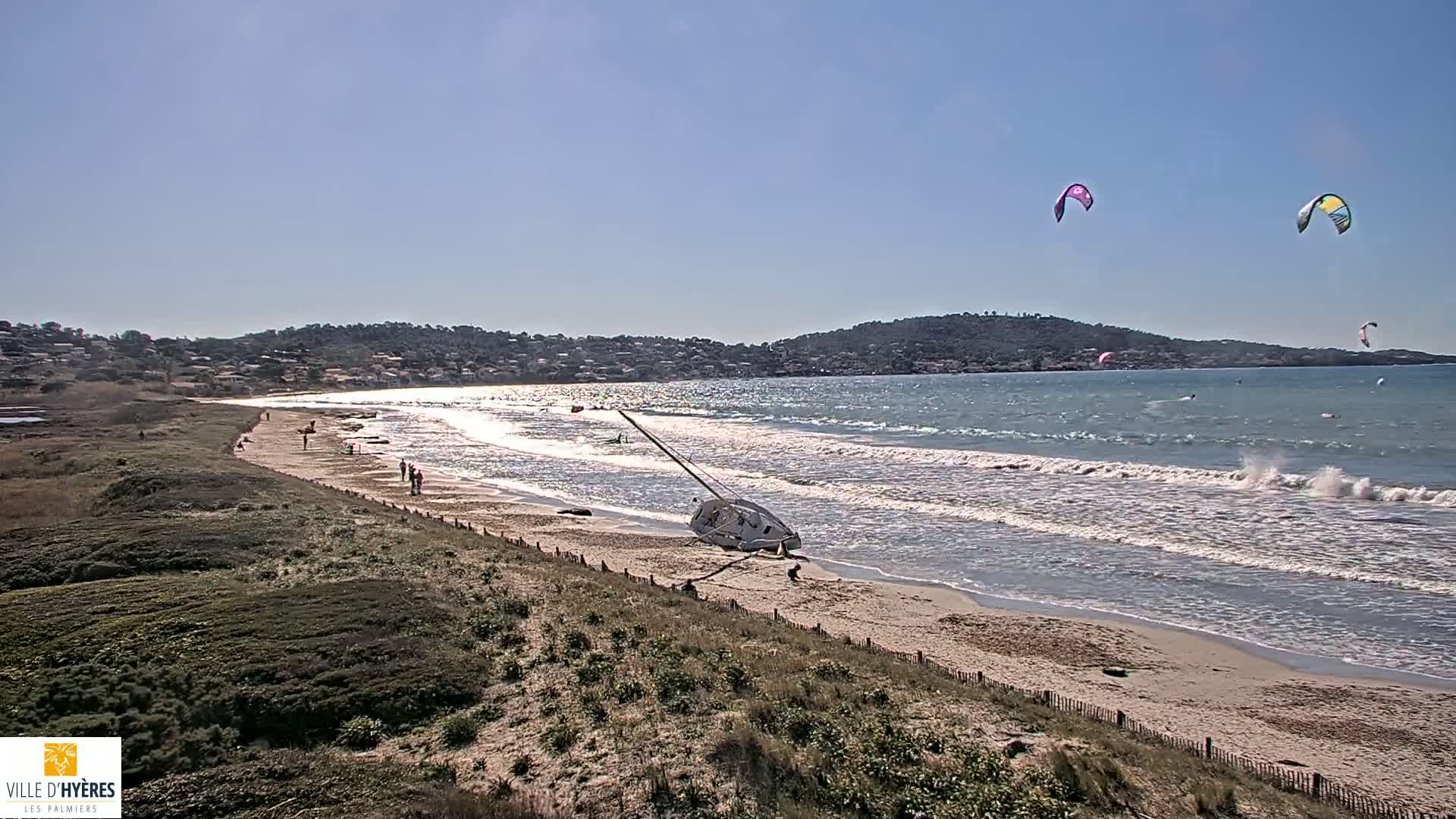 A sandy beach with gentle waves under a sunny sky, a capsized sailboat on the shore, and several kitesurfers in the water.