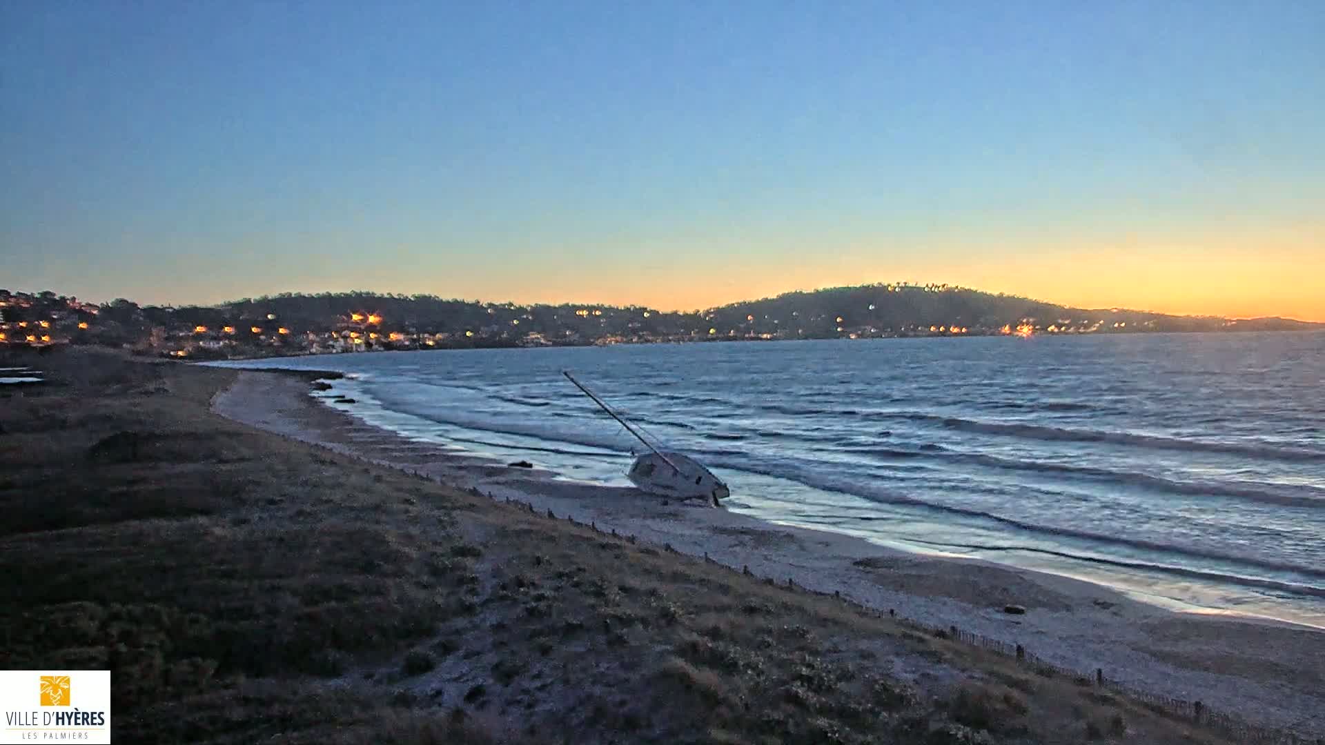 A small sailboat lies on its side on a sandy beach at sunset, with gentle waves lapping the shore under a clear sky.
