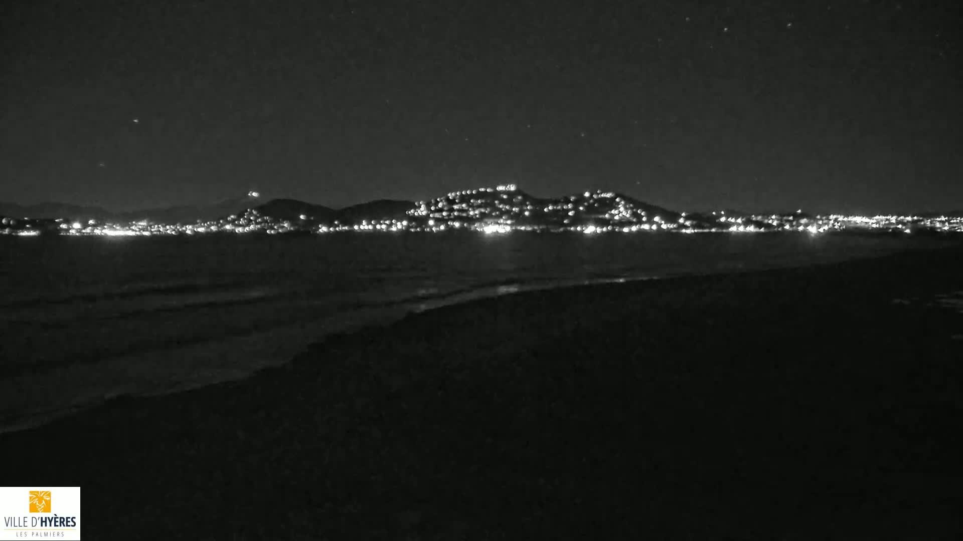 A nighttime view shows a dark beach with gently lapping waves and a distant city illuminated on a hillside across calm water under a clear, starry sky.