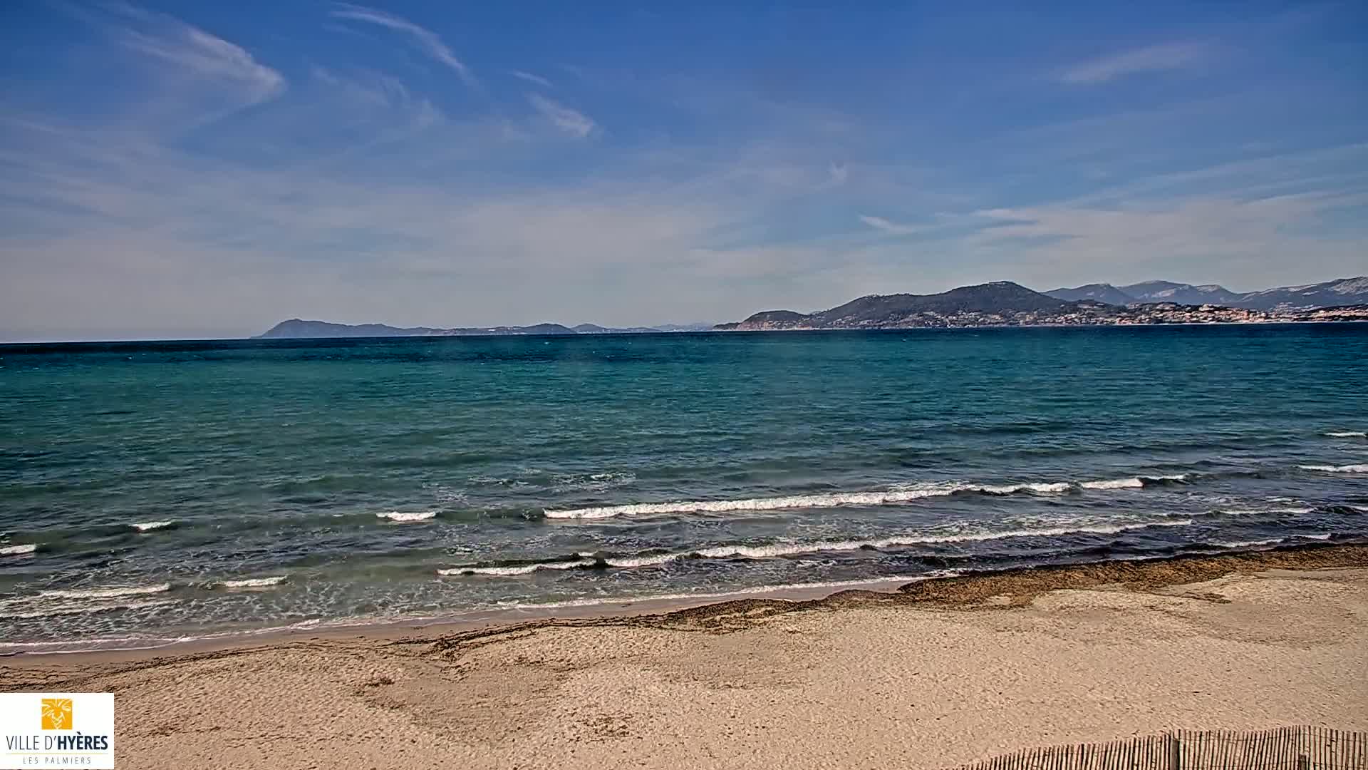 A sandy beach with small waves lapping the shore under a mostly clear, bright sky, with mountains visible in the distance.