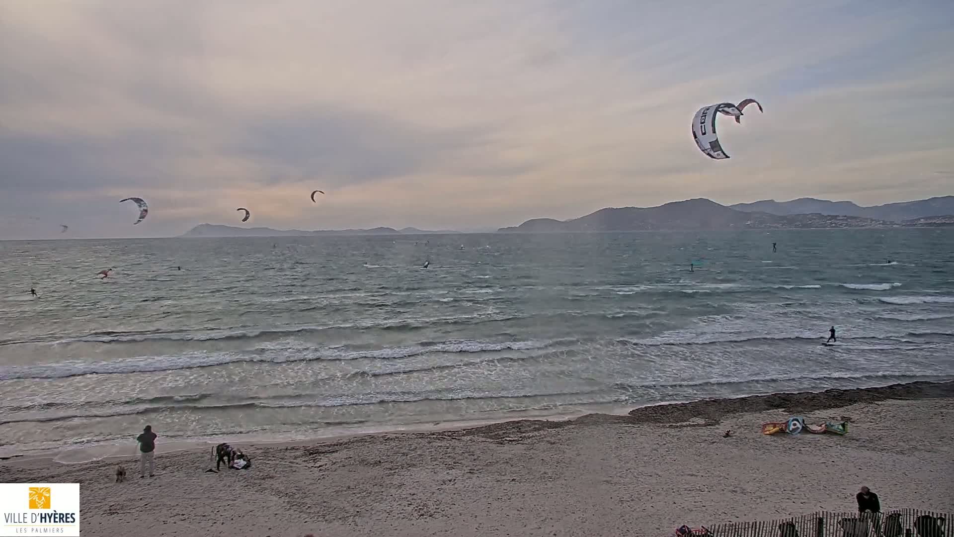 A sandy beach with several people on it overlooks a windy ocean where multiple kite surfers are enjoying the overcast day.