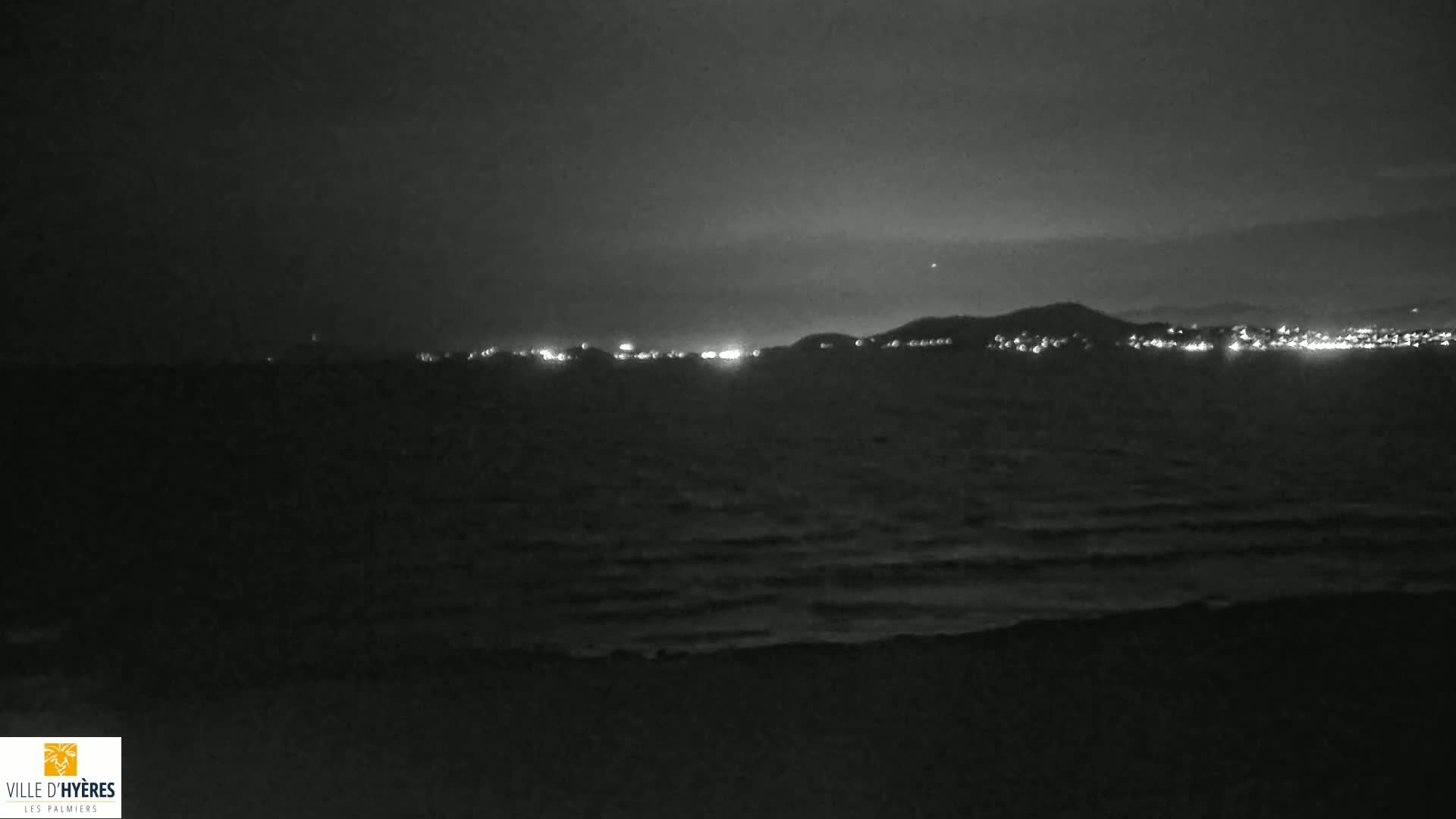 A dark, nighttime coastal scene shows a gently rolling sea with a distant line of lights on the shore.