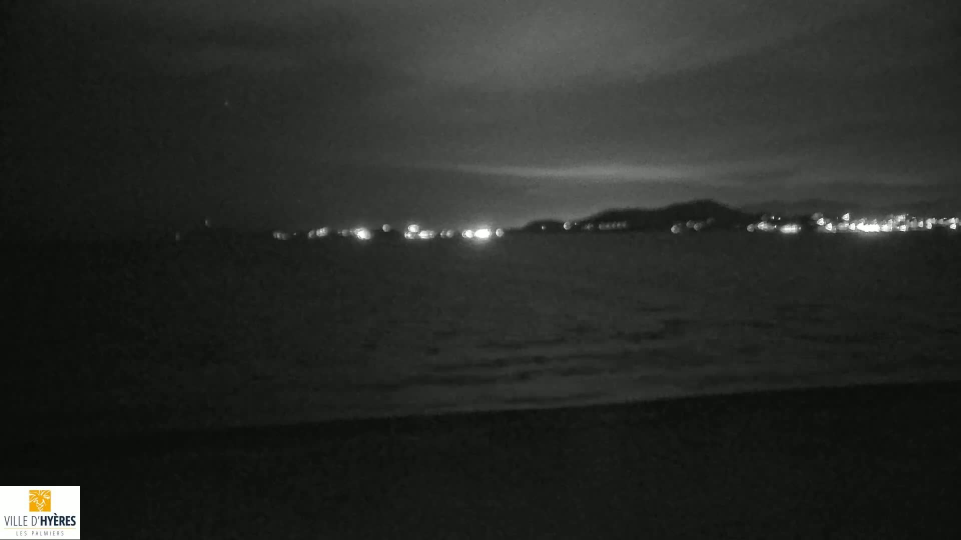 A dark, nighttime beach scene shows a calm body of water with a distant city skyline faintly illuminated.