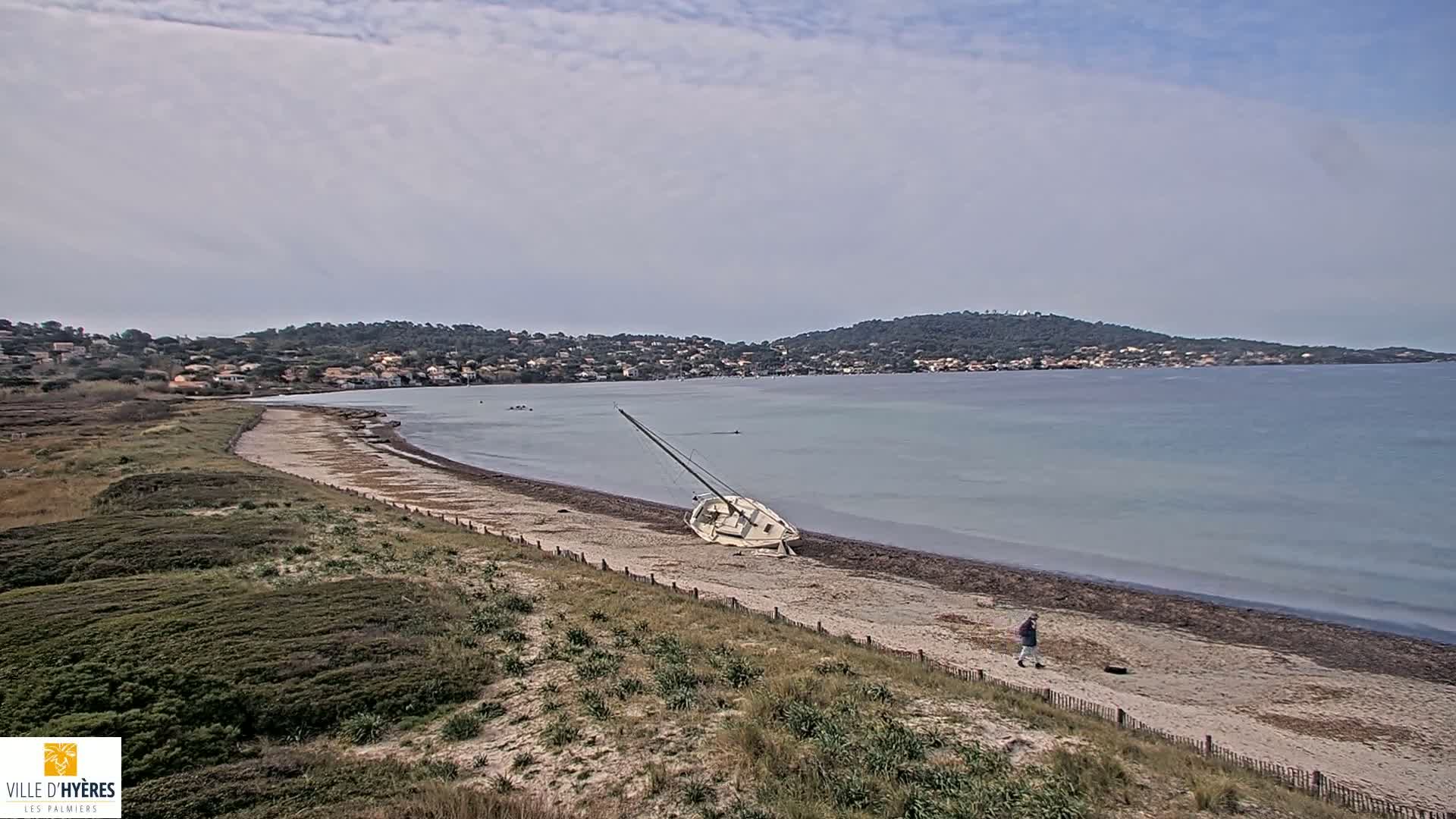 A sailboat lies on its side on a sandy beach, with a person walking nearby, under an overcast sky and calm water in the background.