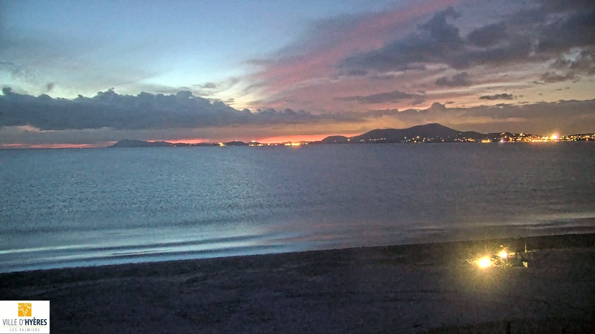 A dark beach at twilight shows a calm sea with a distant city skyline illuminated against a partly cloudy sky with pink and purple hues.