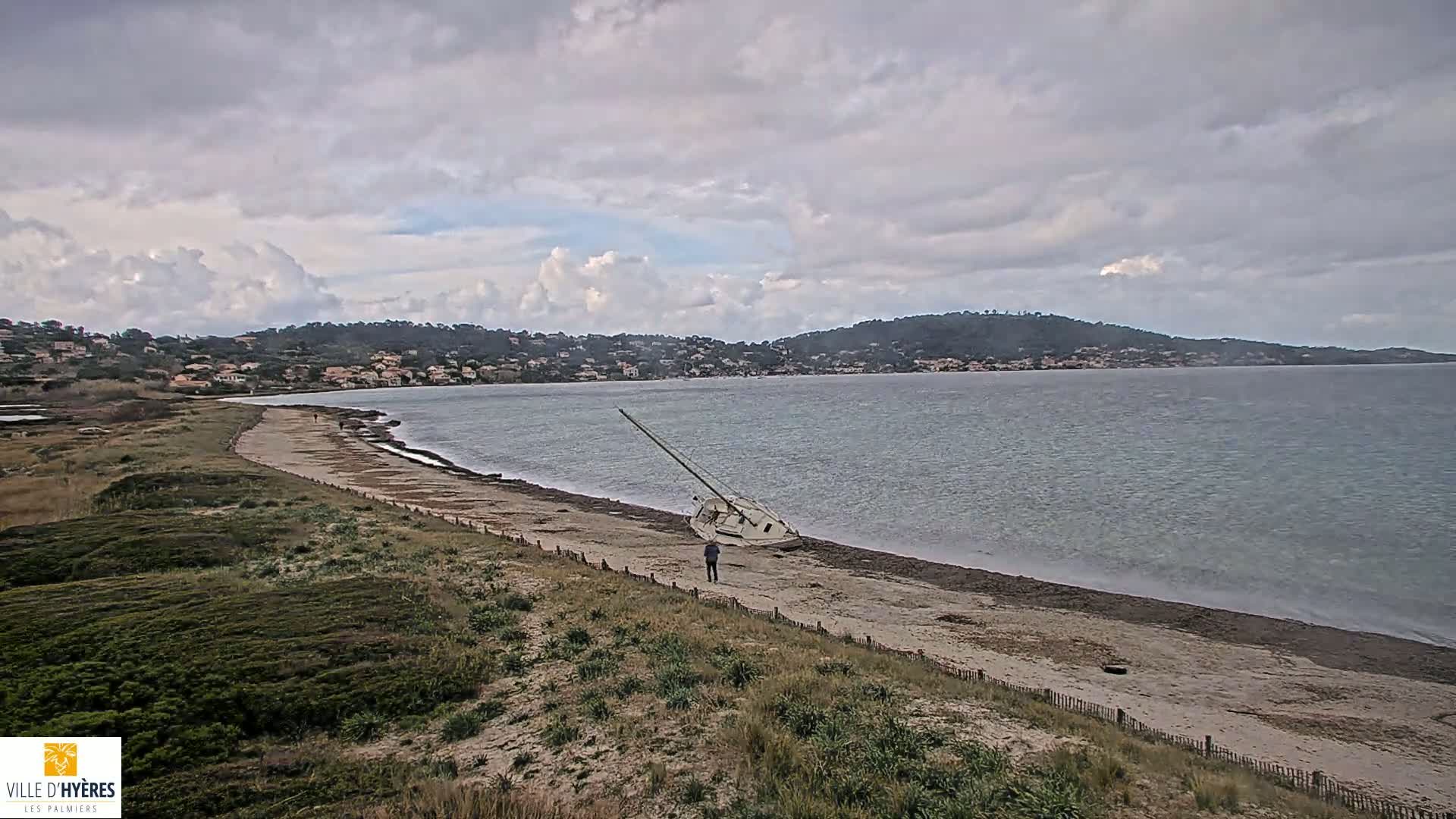 A capsized sailboat lies on a sandy beach near a calm bay under a cloudy sky.