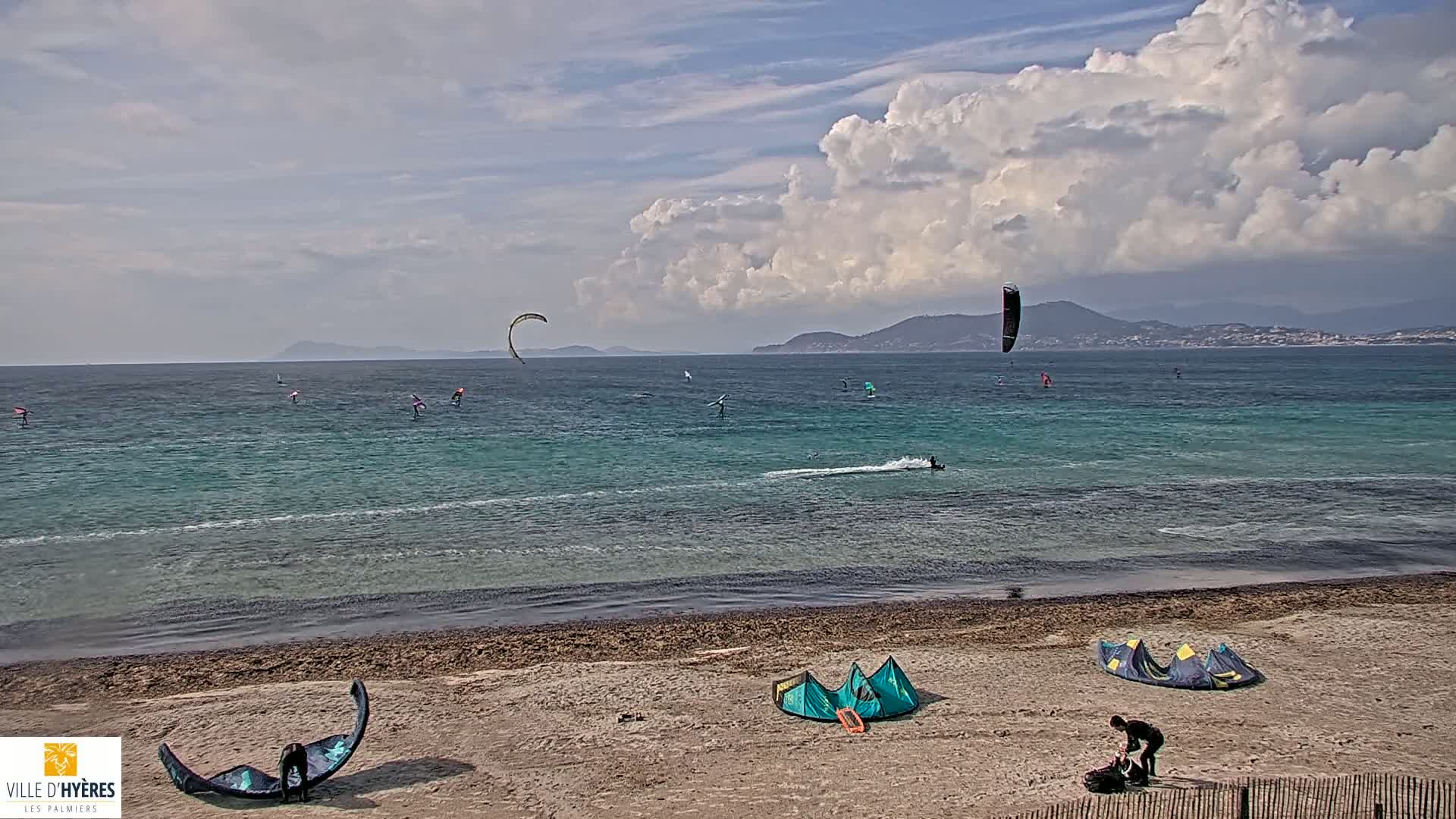 A sandy beach with several kitesurfers and windsurfers in the turquoise water under a partly cloudy sky.