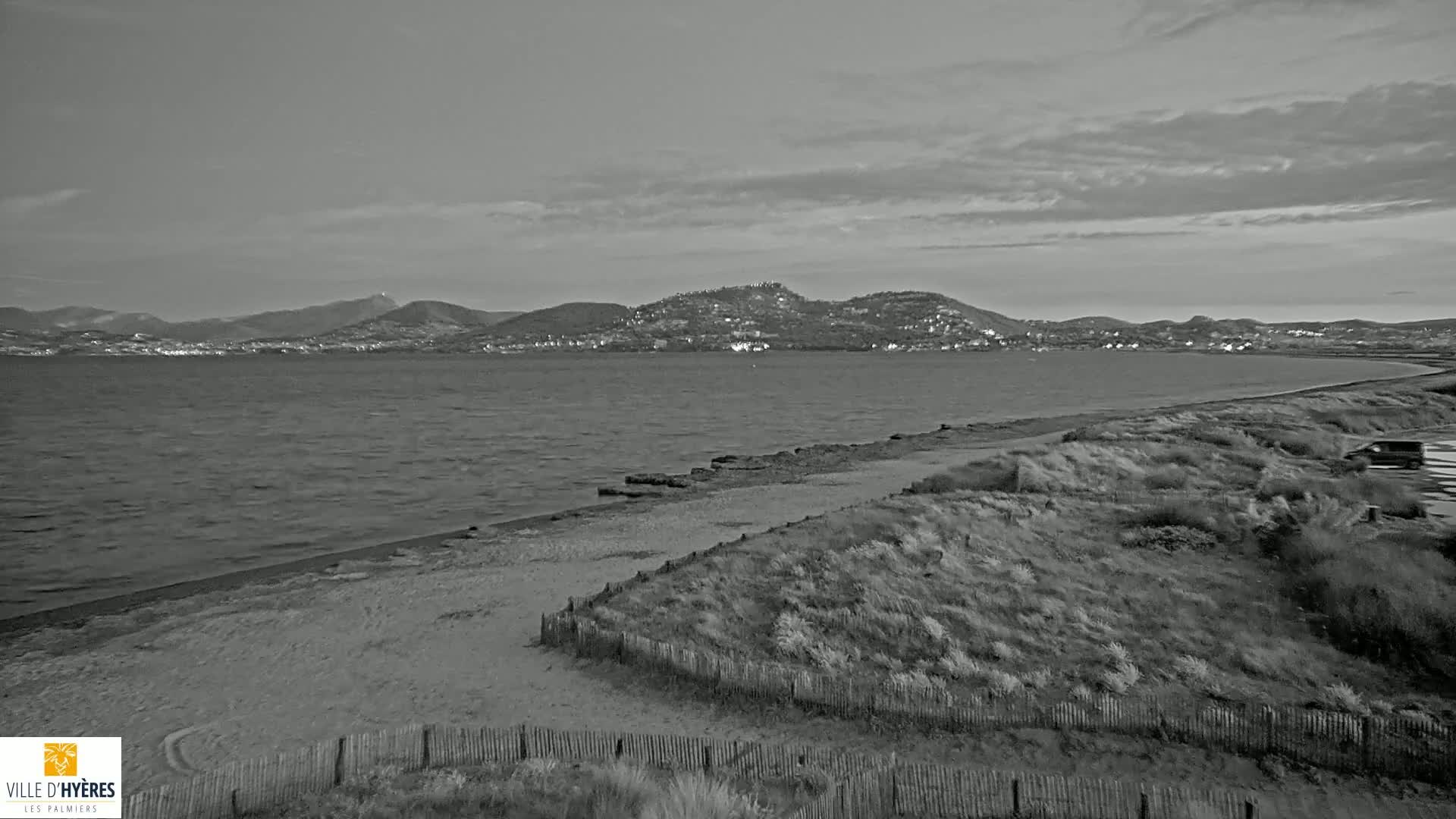 A sandy beach with low dunes and a wooden fence stretches along a calm, gray sea, with a distant mountain range under a cloudy sky.