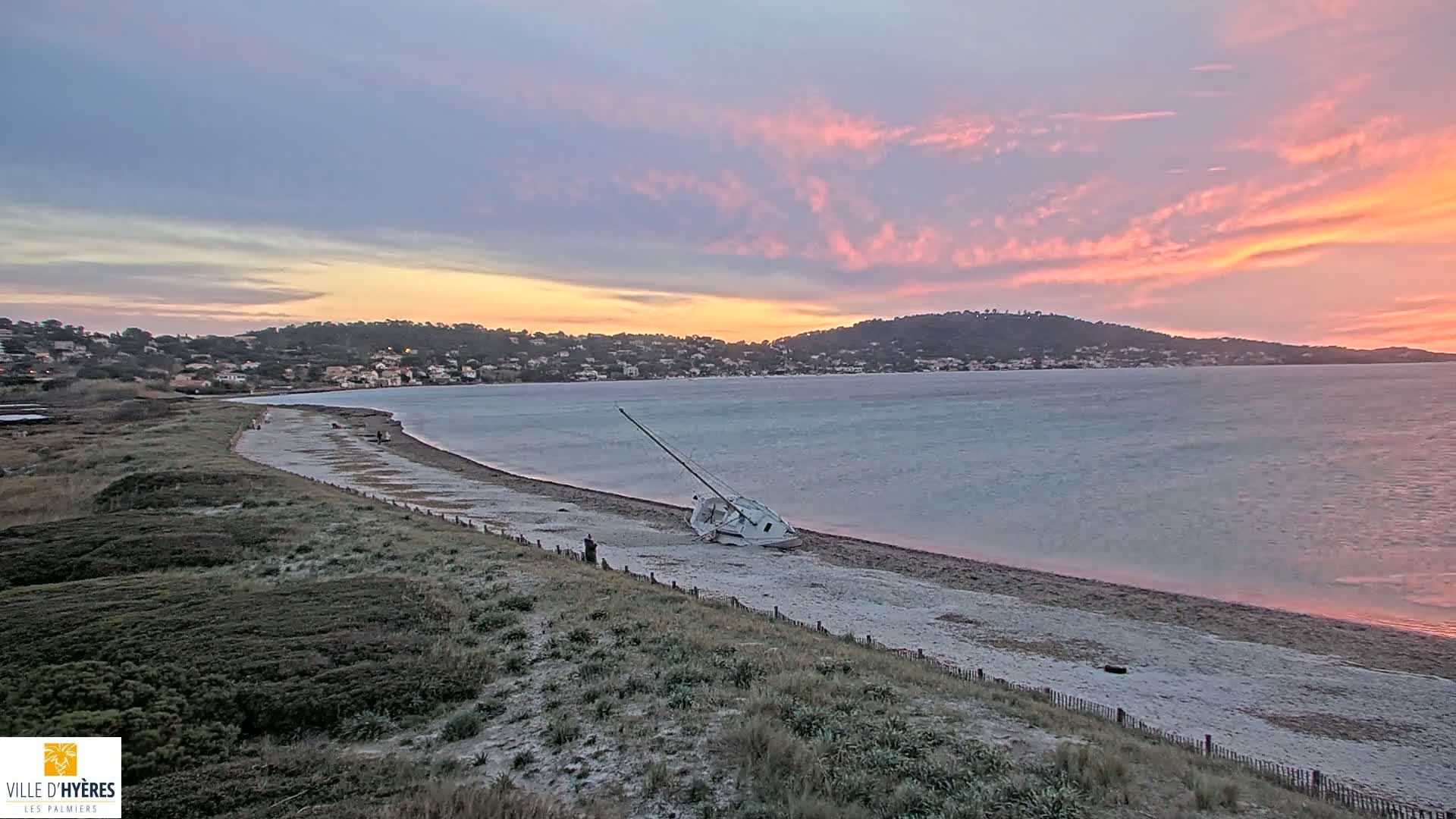 A sailboat lies on its side on a sandy beach at sunset under a pink and purple sky.
