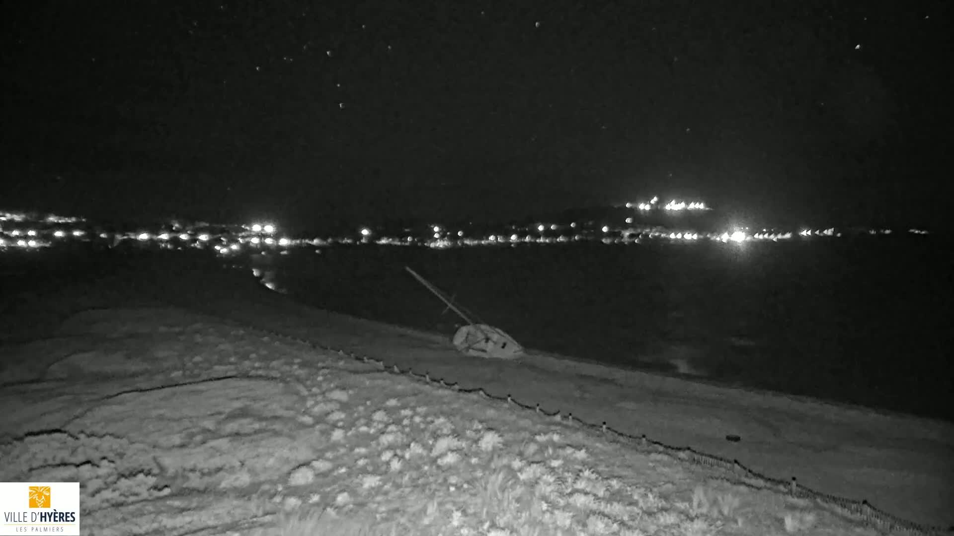 A beached boat lies on a dark, sandy shore at night under a starry sky, with distant city lights visible across the water.