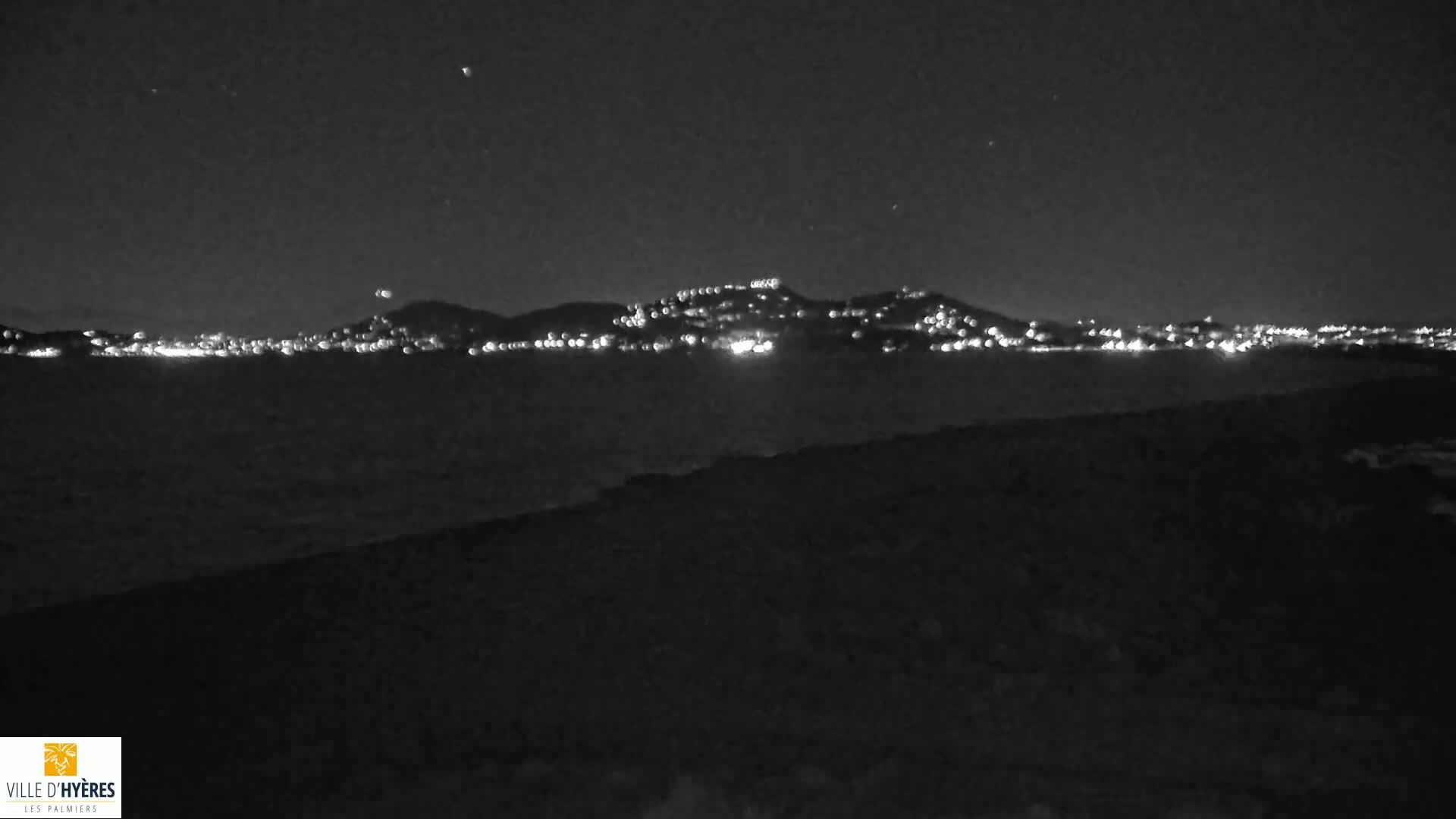A nighttime view shows a dark, hilly coastline with numerous lights illuminating a city or town in the distance under a clear, starry sky.