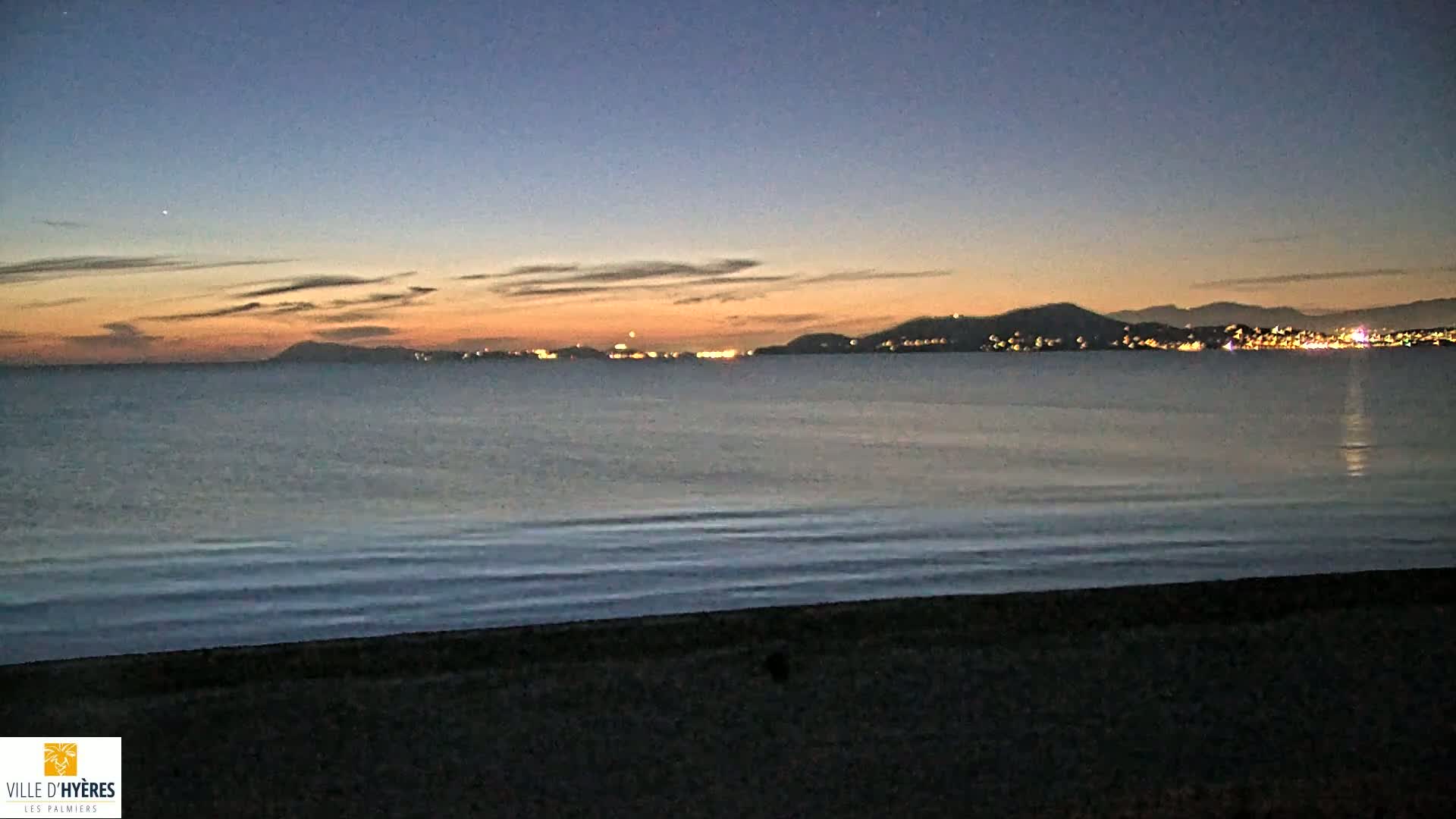 A dark beach at twilight overlooks a calm sea with a distant city skyline illuminated at night.