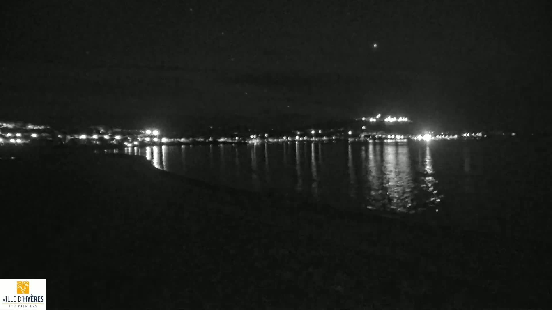 A nighttime view of a calm body of water reflecting the lights of a distant town under a clear, starry sky.