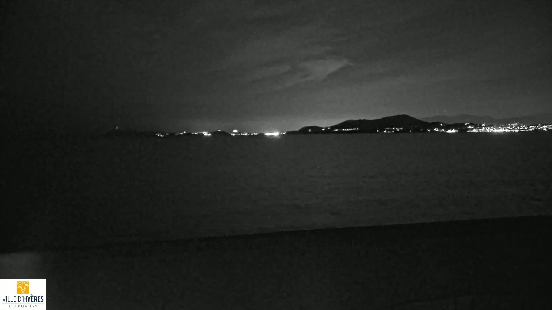 A nighttime view of a dark body of water with faintly lit hills and a cityscape in the distance under a cloudy sky.
