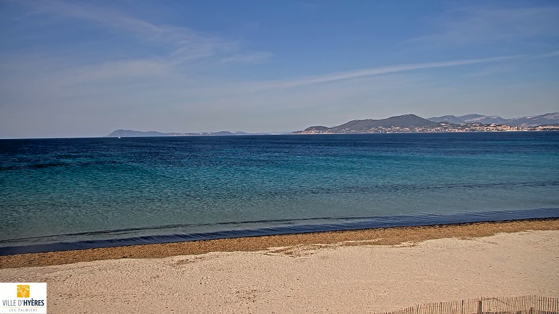 A sandy beach with calm, turquoise water stretches to a distant mountain range under a clear, sunny sky.