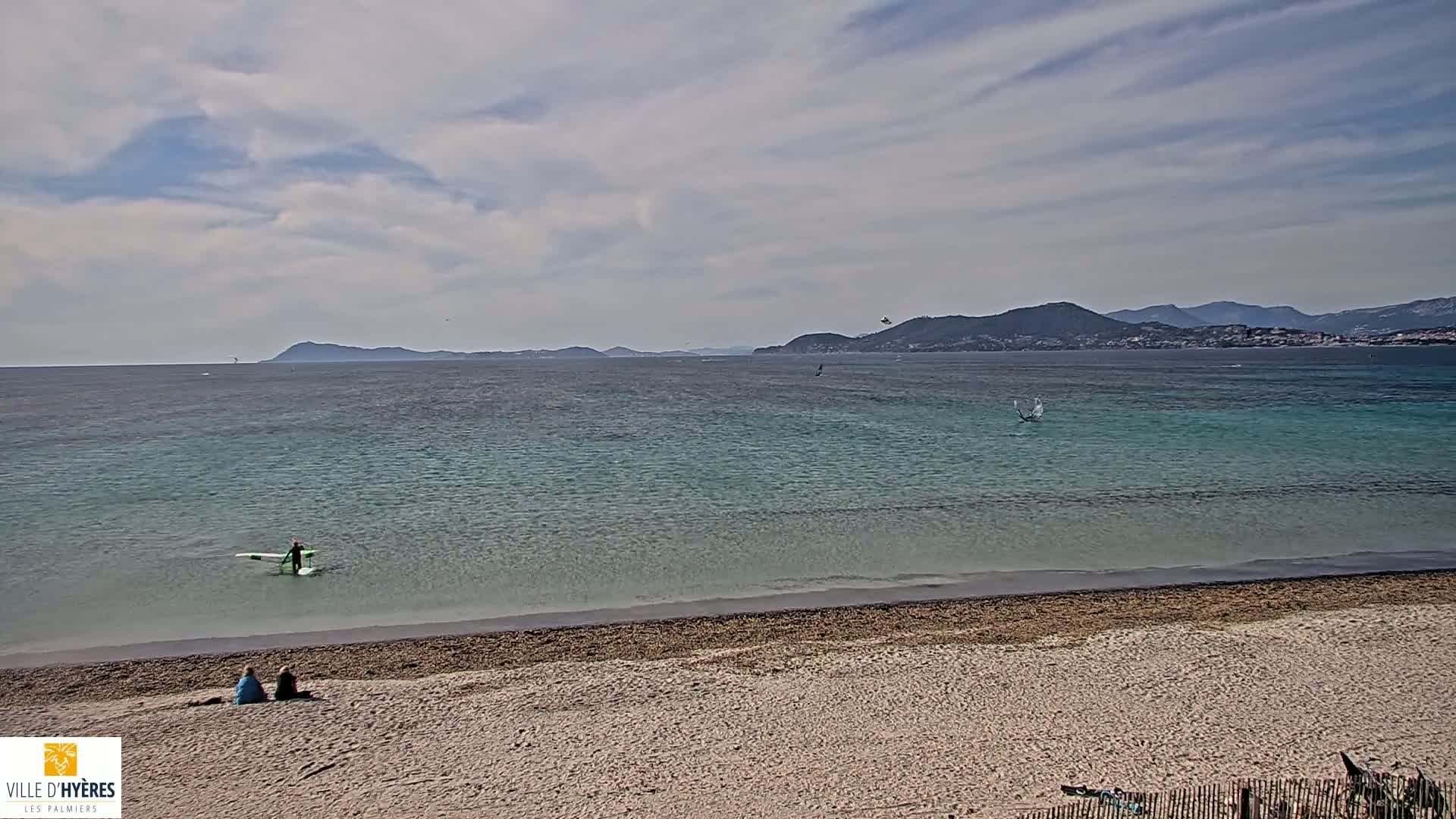 A sandy beach with two people sitting on it overlooks a calm, light-blue ocean with a person stand-up paddleboarding in the distance under a partly cloudy sky.