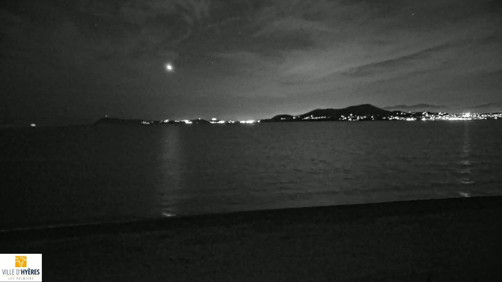 A nighttime view shows a dark body of water with a distant, dimly lit shoreline under a cloudy sky.