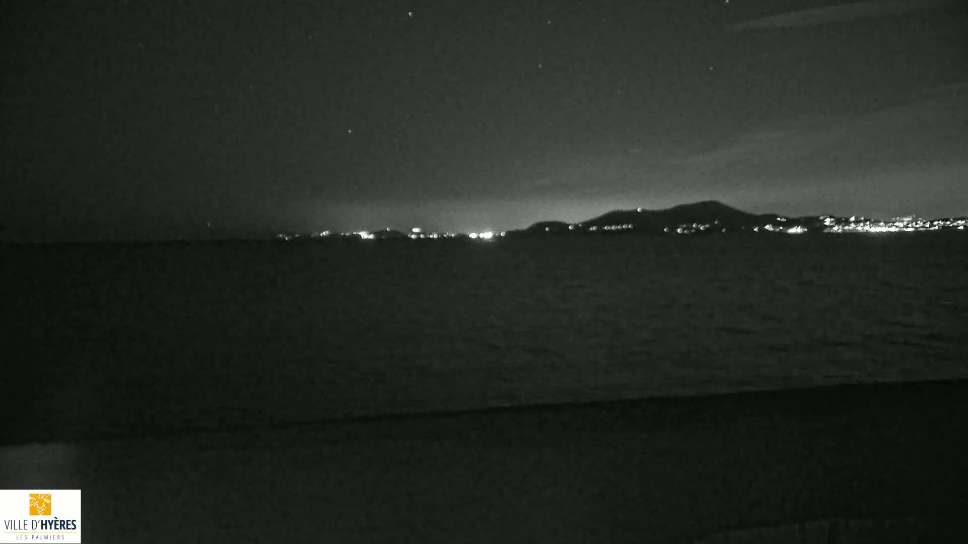 A nighttime, dark view of a calm body of water with a distant, illuminated city skyline under a clear, starry sky.