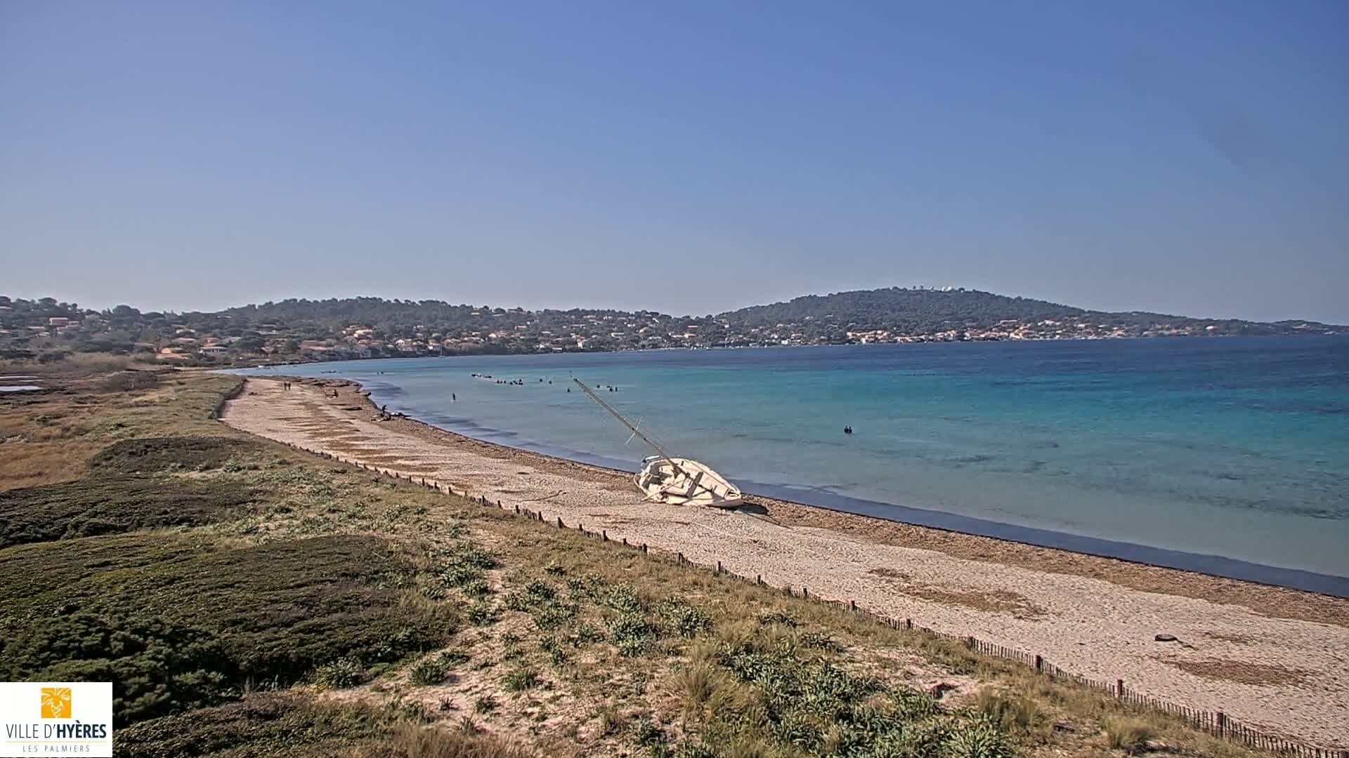 A sandy beach with a capsized sailboat lies on the shore next to clear, teal water under a sunny sky, with a town visible in the distance across the bay.