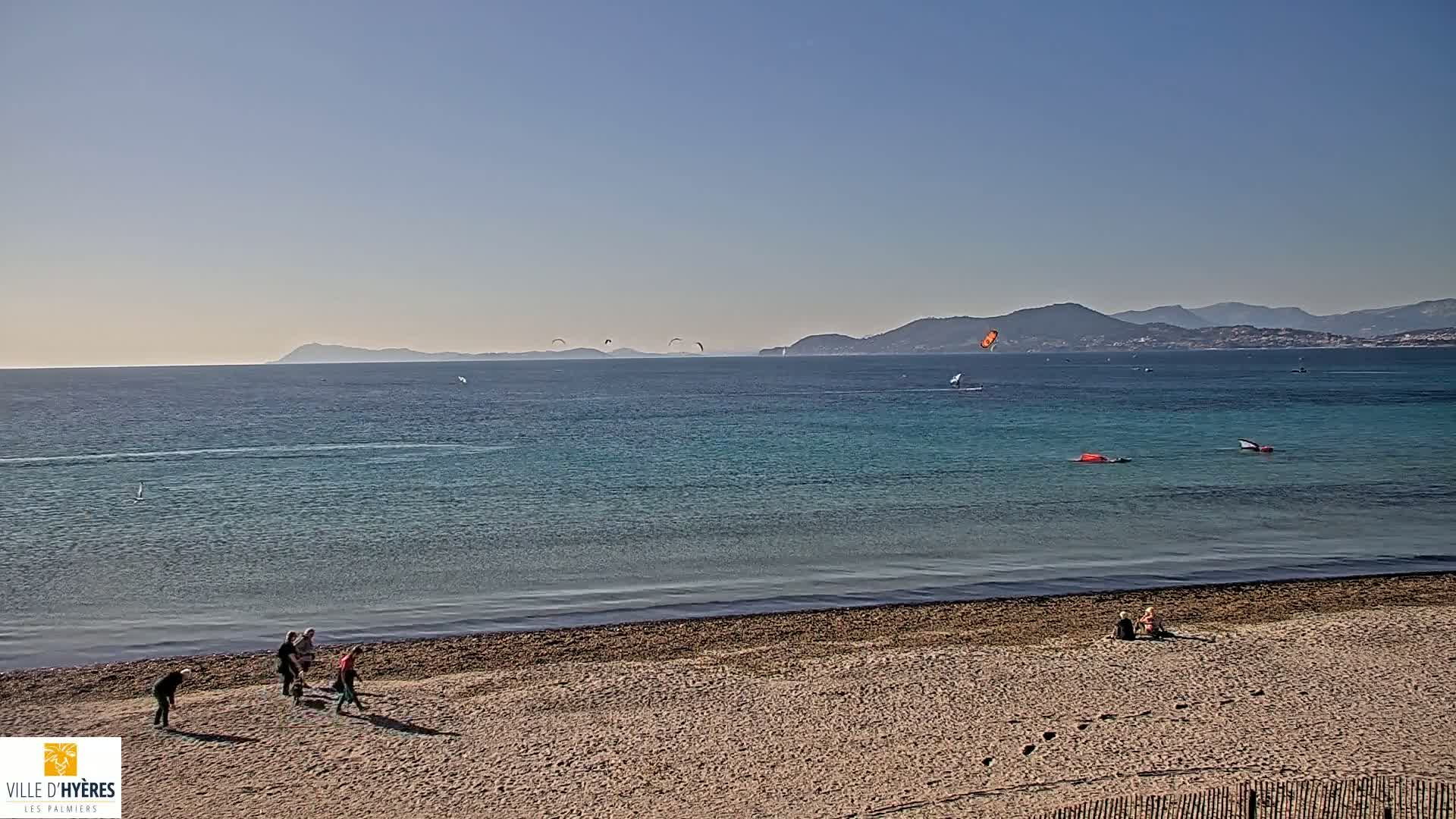 A sandy beach with several people walking near the water's edge, overlooks a calm, teal sea with sailboats and kitesurfers visible in the distance under a clear sky.